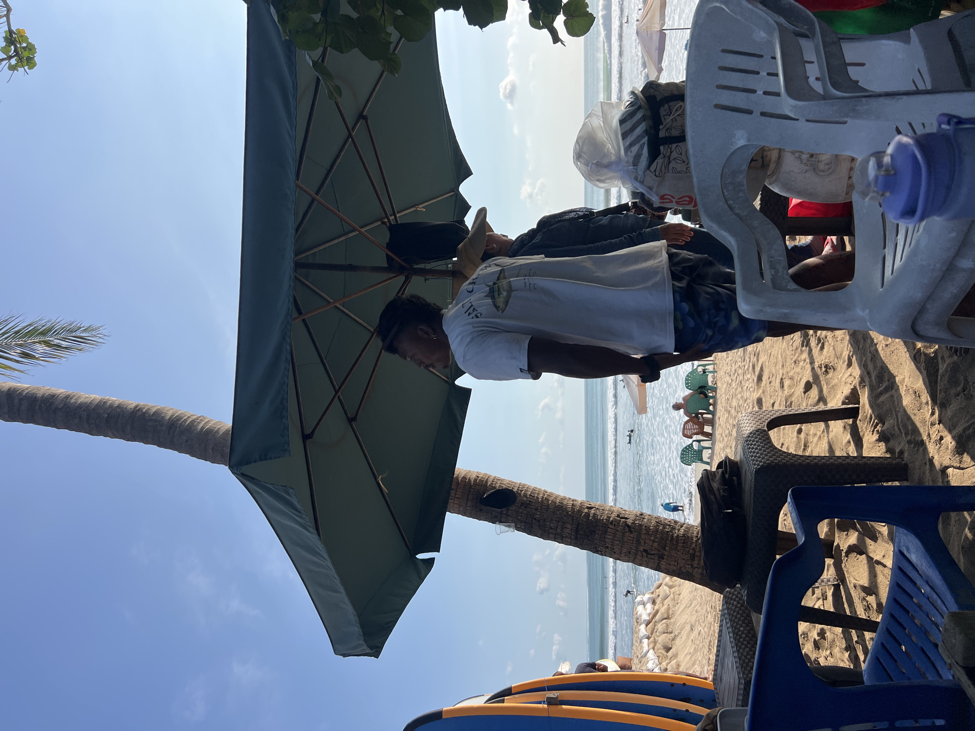 On a sunny day at Kuta Beach, Indonesia, several people are enjoying the shore. In the foreground, a blue plastic chair is positioned on the sandy beach, with a gray plastic chair stacked on top of it. To the right of these chairs, a large green umbrella casts a shadow over the scene. Beneath the umbrella, two individuals stand, one wearing a white t-shirt with a graphic and patterned shorts, and the other wearing a denim jacket and a straw hat. A palm tree trunk stands next to them. In the background, the ocean stretches out with small waves, and a few surfers can be seen in the water. The beach is lined with sandbags and beach chairs, with a glimpse of other umbrellas and a few more people sitting in the distance. Surfboards are leaning against a wall on the left side of the image.