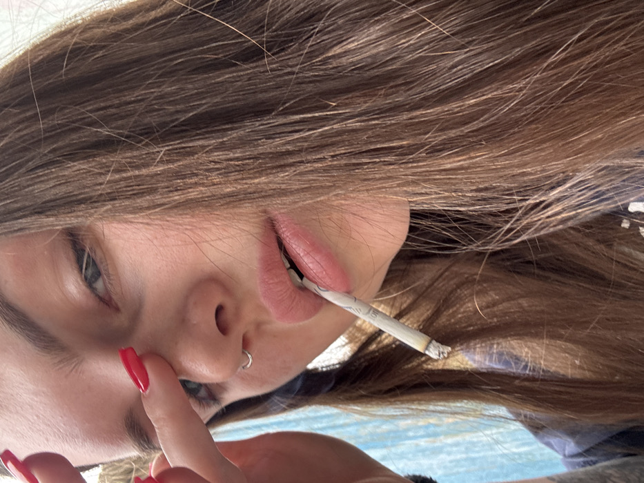 The image displays a close-up, sideways portrait of a young person, likely taken from a low angle or with the camera rotated. The subject's face is positioned in the lower-left to central area of the frame.
The person has long, straight, light brown hair with lighter streaks, which spreads across the top and right side of the image, obscuring much of the background. Their visible left eye, a light color such as blue or green, is looking directly towards the viewer. A small, circular silver nose ring is present in their left nostril.
The subject's lips are full and tinted with a pale pink color. They are slightly parted, holding a hand-rolled cigarette or joint. The wrapper of the rolled item is white, with faint, repeating patterns or text visible along its length. The tip of the item shows a small amount of gray ash, indicating it is lit or has been recently smoked.
A hand with long, almond-shaped fingernails, painted in a vibrant red, is visible in the lower-left corner, with one finger gently resting near the cheek or temple area.
The background behind the person's hair is largely out of focus, revealing a blend of light blue and white tones, suggesting an outdoor setting like a sky or a distant, blurred surface. The overall activity depicted is the subject engaging in smoking while looking directly at the camera.