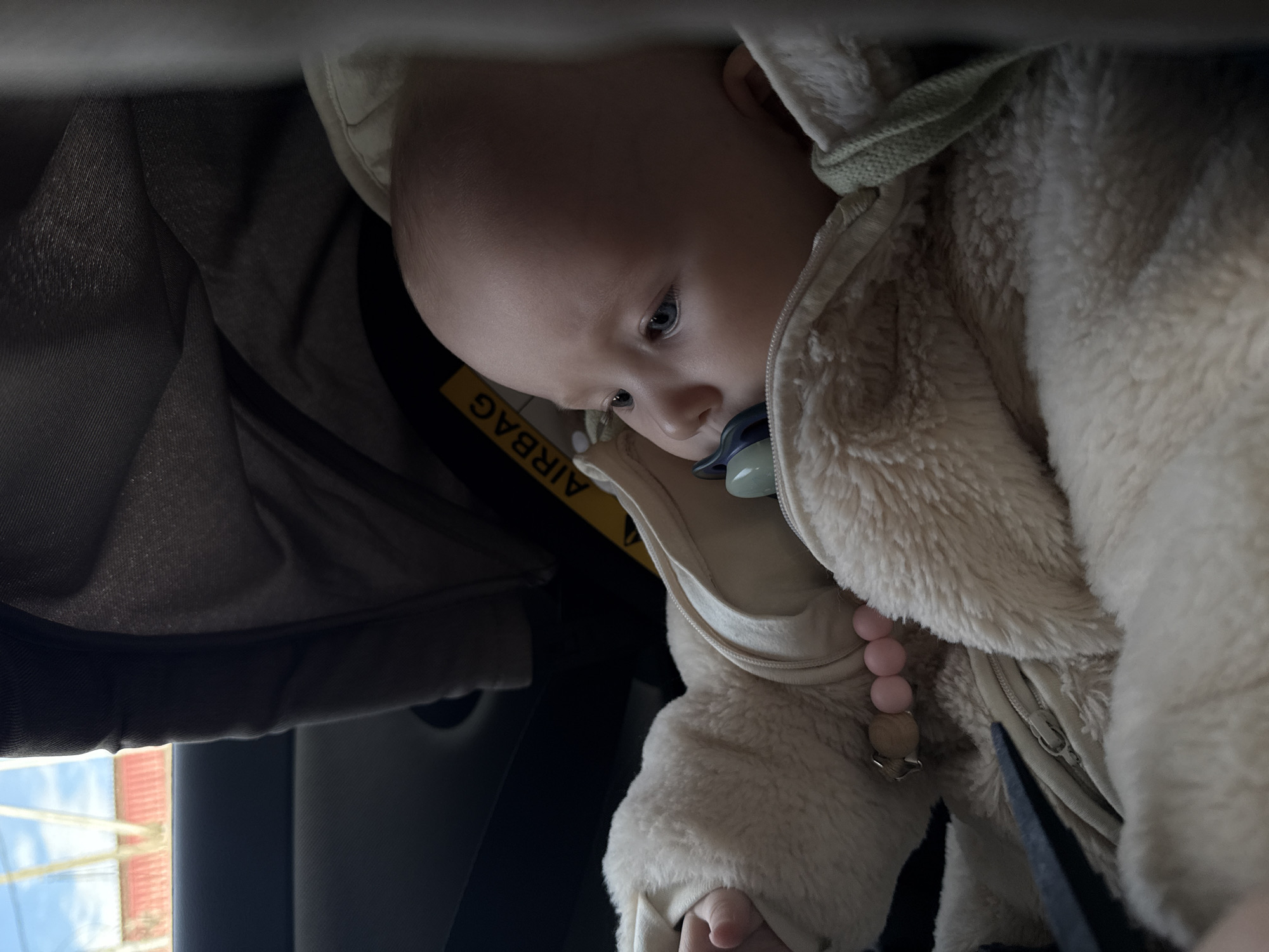 A baby with blue eyes is looking down and to the left, with a pacifier in their mouth. The baby is wearing a fuzzy cream-colored hooded jacket with a visible zipper and pink and wooden bead details. The background appears to be the interior of a vehicle, possibly a car seat, with a yellow tag that reads "AIRBAG". Outside the car window, a red building is visible. Given the information provided, it's assumed the scene is within the city of Yakutsk, Russia.