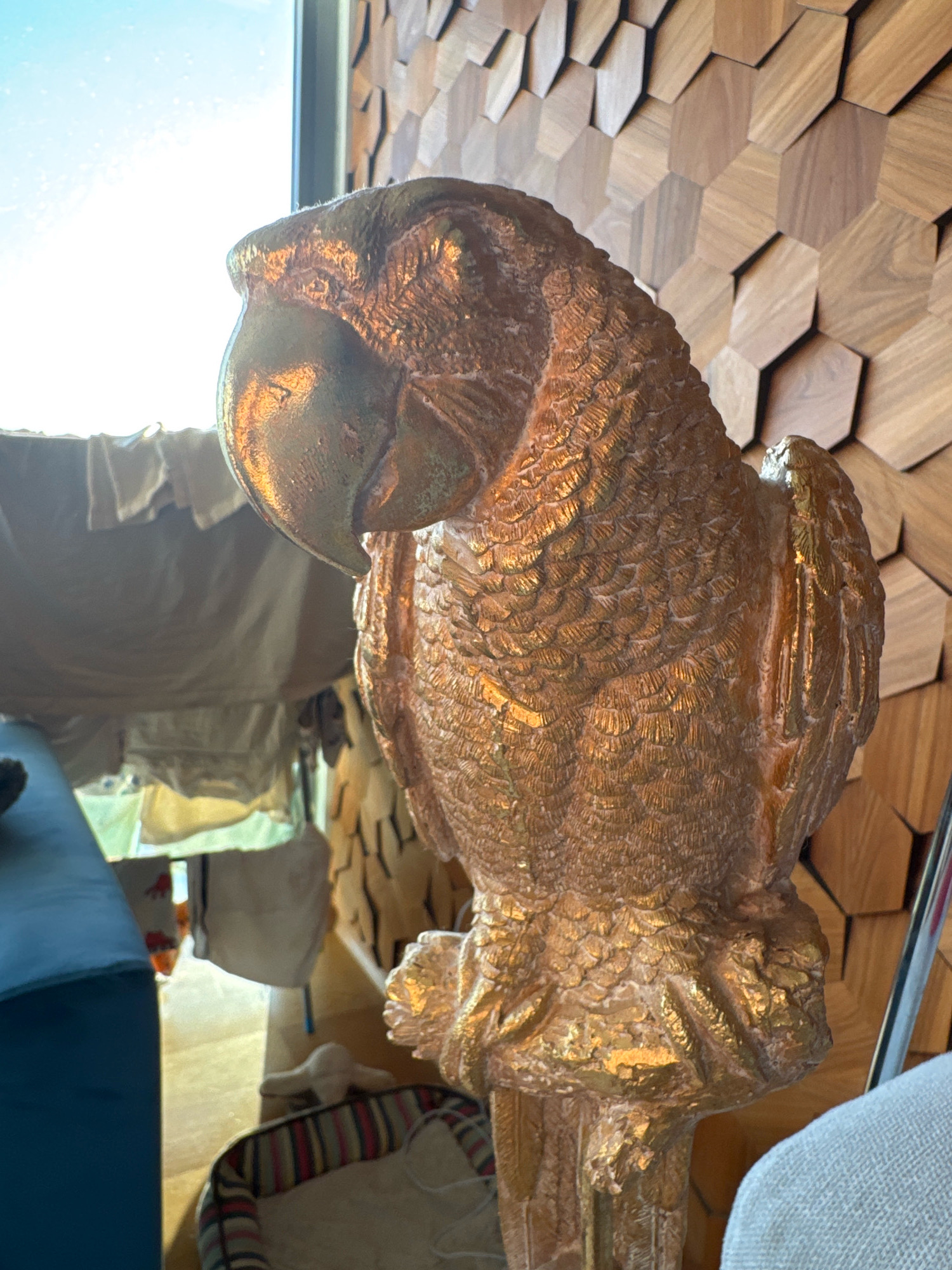 A golden statue of a parrot is positioned centrally in the foreground, angled to the left, facing away from the camera. The parrot statue is highly textured, with individual feathers clearly defined, and its surface reflects light, giving it a metallic sheen. It stands on a base, which is also part of the statue.
Behind the parrot statue, a wall with a geometric, honeycomb-like pattern made of wood panels serves as the backdrop. To the left of the statue, sunlight streams in from a window, casting a bright light and creating a slightly washed-out effect in that area. Partially visible beneath the window are hanging textiles, possibly clothing or curtains, and a blue surface, likely a piece of furniture. Further into the background, a pet bed with colorful stripes is visible, containing a white object, possibly a toy.
The scene appears to be indoors, in a room with a distinct decorative wall. The lighting suggests it is daytime. There are no people visible in the image. The environment is clean and well-lit, with a focus on the ornate parrot statue. This scene could be located in a residential or commercial space in Limassol, Cyprus, given the request.