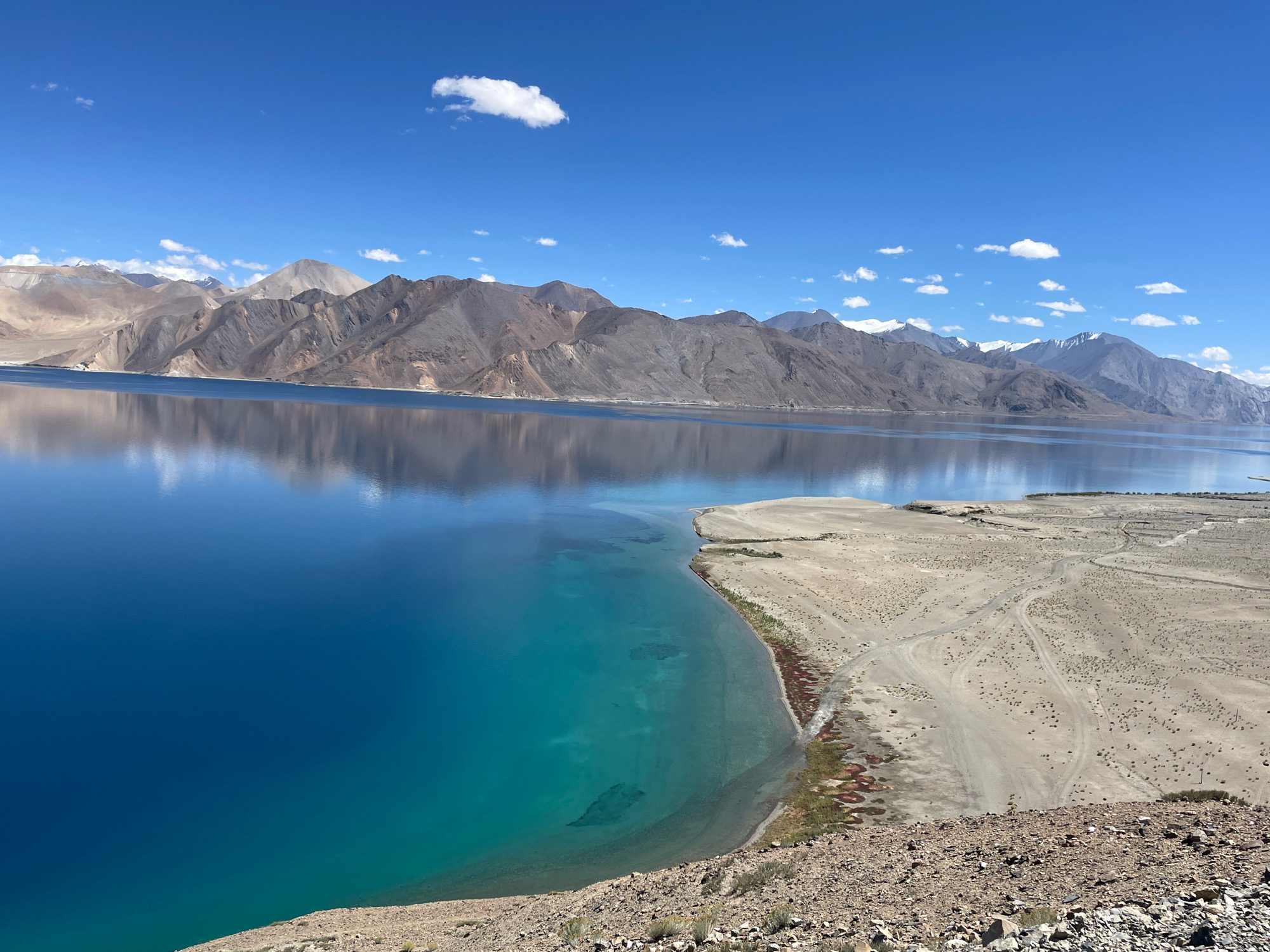 An aerial, high-angle, wide shot shows the vast expanse of Pangong Tso lake in India. The lake's surface is a brilliant, deep blue that transitions to a lighter, turquoise hue closer to the shore. The clear water allows visibility of the lakebed in the shallower areas. A sandy, arid shoreline stretches out, marked by faint tire tracks from vehicles. The background is dominated by a mountain range with rugged, barren slopes. The sky above is a vibrant blue, dotted with a few wispy white clouds. The reflection of the mountains and sky is clearly visible on the placid surface of the lake, creating a symmetrical effect. The foreground is composed of rocky, barren terrain, suggesting a high-altitude, desert-like environment.