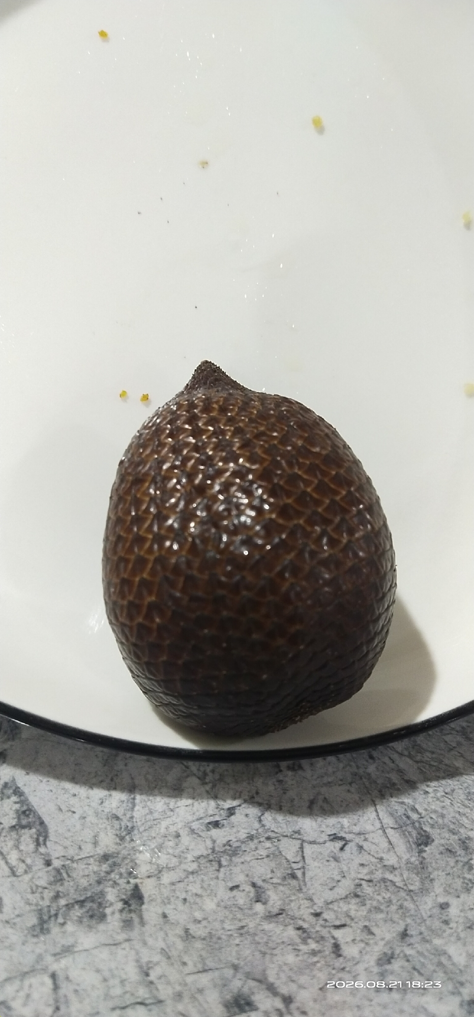 A single salak fruit, also known as snake fruit, is centered on a white plate with a black rim. The fruit has a rough, scaly, dark brown skin and tapers to a point at the top. Scattered across the white plate are a few tiny yellow specks, likely fragments of food. The plate rests on a gray, marbled surface. The image captures a close-up view of the fruit and plate. The timestamp "2026.08.21 18:23" is visible in the bottom right corner. This image does not contain enough information to determine its specific location within Matabu, Indonesia, or to identify any people or actions.