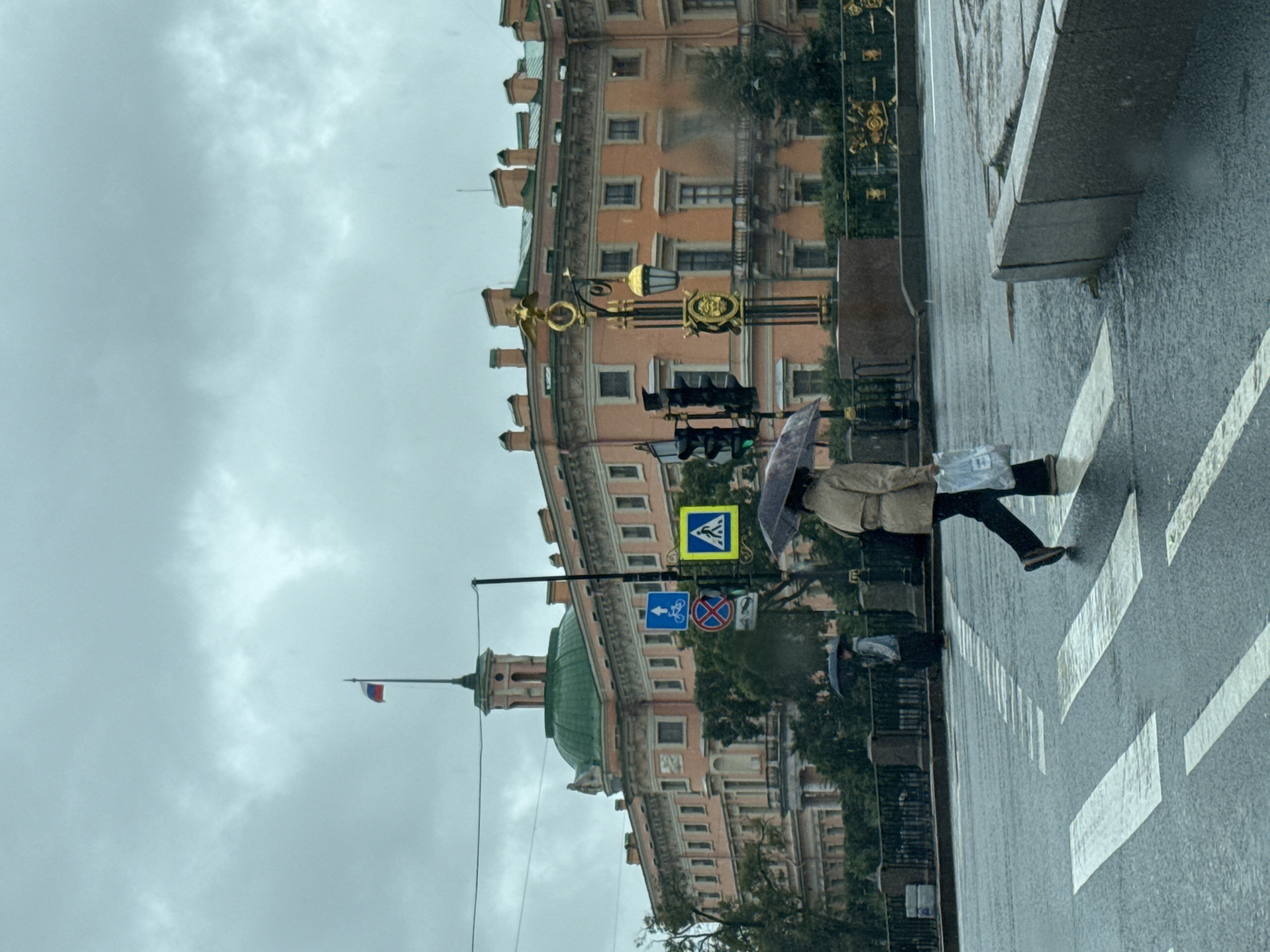 The image depicts a street scene in Saint Petersburg, Russia, with a view of the Mikhailovsky Castle in the background. The weather appears to be overcast and rainy, with the road and crosswalk wet. A pedestrian is crossing the street, holding an umbrella and carrying a plastic bag. Another pedestrian with an umbrella is visible further down the street. Traffic lights and street signs are also present. The Mikhailovsky Castle is a prominent architectural landmark, characterized by its orange facade, green dome, and ornate detailing. The overall atmosphere is one of a typical, somewhat gloomy, urban day in Saint Petersburg.