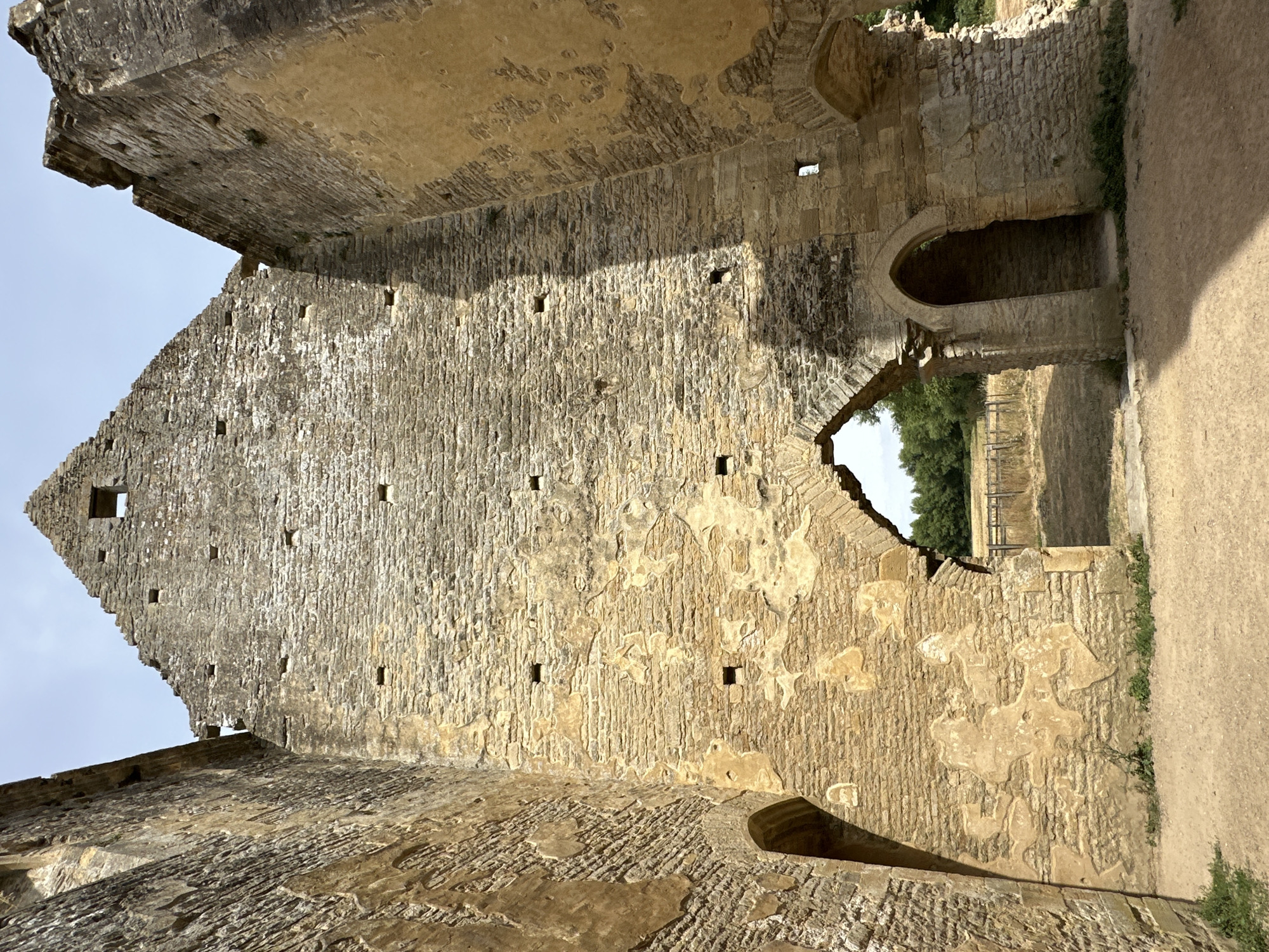The image shows the interior of a stone ruin with a triangular-shaped roofline against a pale sky. The walls are made of rough-hewn, tan-colored stones, with some areas showing patches of wear and missing mortar. Two large arched openings are visible on either side of the central facade, framing a view of dry grass, a wooden fence, and trees in the distance. A smaller arched doorway is situated within the central section, leading into a dark interior. The ground in front of the ruin is covered in dirt and small patches of green vegetation. The scene is bathed in sunlight, casting shadows on the stone surfaces.