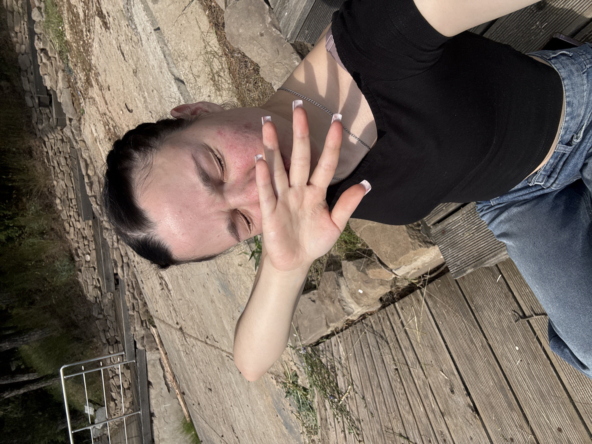 A young woman with dark hair braided and pulled back sits on a wooden bench outdoors, covering her face with her left hand. Her eyes are closed, and her facial skin appears flushed. She is wearing a black cropped t-shirt and blue jeans. Behind her, a stone embankment with steps and a metal railing leads upwards towards trees. The ground is a mix of concrete and wooden planks. There is no indication in the image that this scene is located in Pozdyshki, Russia.