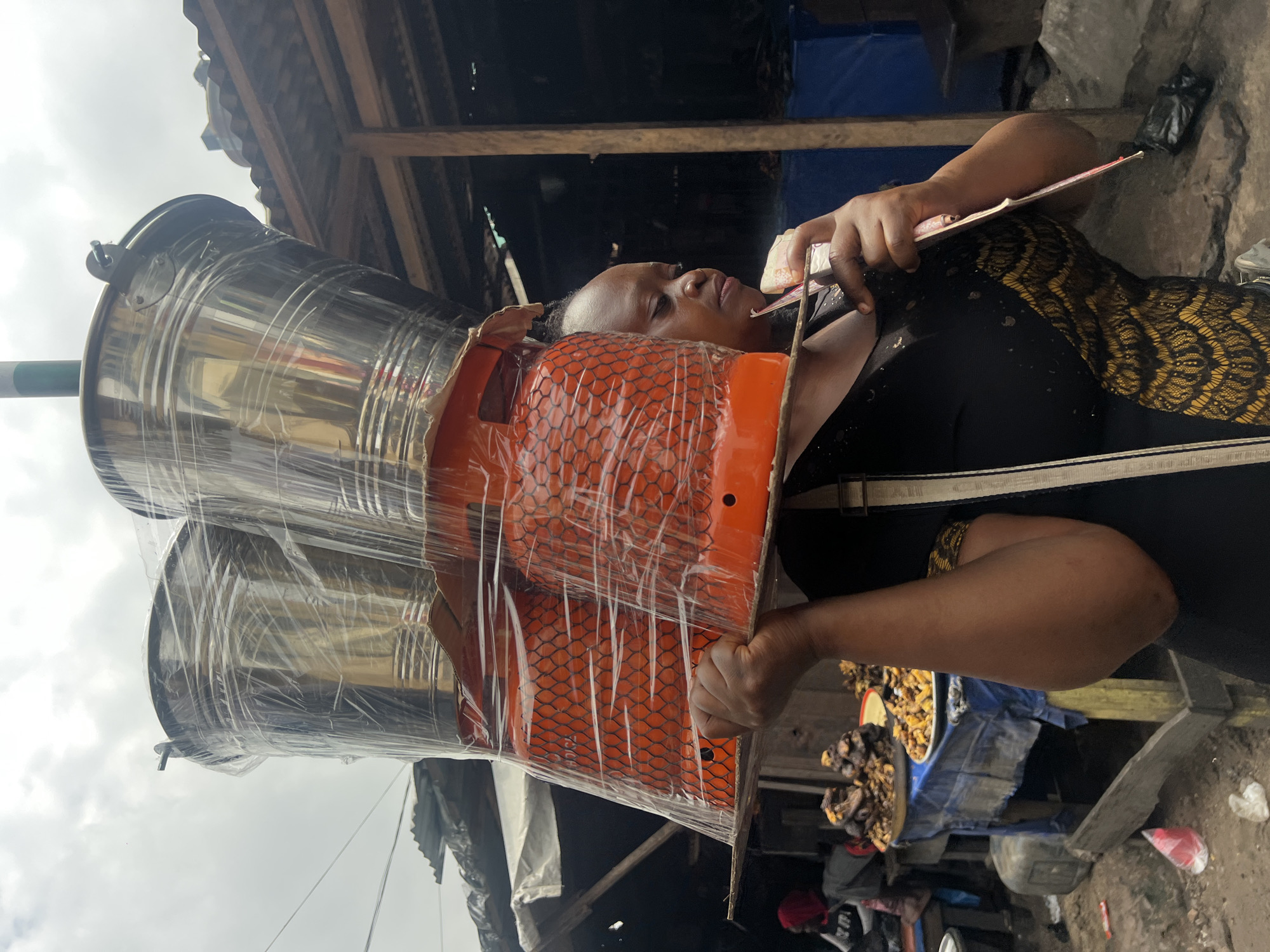 In Nkpor, Nigeria, a woman balances two orange gas cylinders and two metal buckets on her head. The cylinders are wrapped in a white net and clear plastic, as are the buckets. She wears a black dress with a gold and black patterned accent on the side and a white strap from a bag is visible over her shoulder. She holds a pink object, possibly a piece of paper or a fan, in her right hand. The scene appears to be in an outdoor market, with stalls and goods visible in the background, including what looks like food on tables covered with blue tarpaulins. The ground is uneven and appears to be dirt.