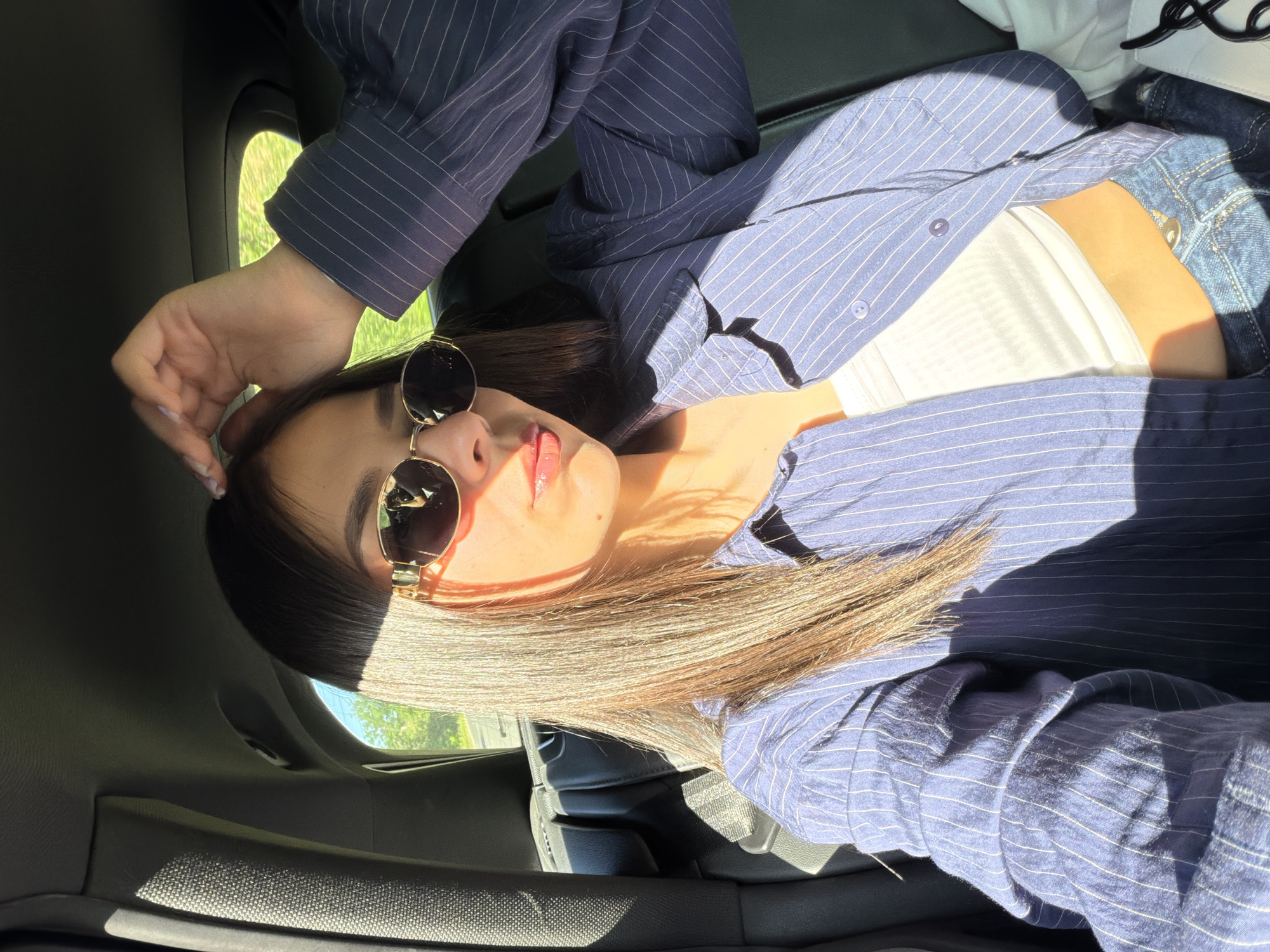 A woman is reclining in what appears to be the passenger seat of a vehicle. She is wearing a striped blue button-down shirt over a white top and denim jeans. She is also wearing sunglasses. Sunlight streams into the car, creating shadows across her face and clothing. The exterior, visible through the window, suggests an urban or suburban setting with trees and possibly buildings. The location in Dryanovo, Bulgaria is not clearly discernible from the image.