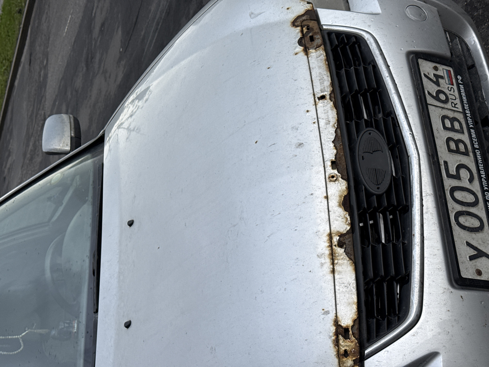 The image shows the front of a silver Lada car with significant rust damage along the top edge of the grille. The car is parked on an asphalt surface, possibly a street or parking lot, with a grassy area visible in the background on the right. The license plate is clearly visible and reads "Y 005 BB 64 RUS". There are no people or other vehicles prominently featured in the image. The overall impression is of a used vehicle in a state of disrepair.