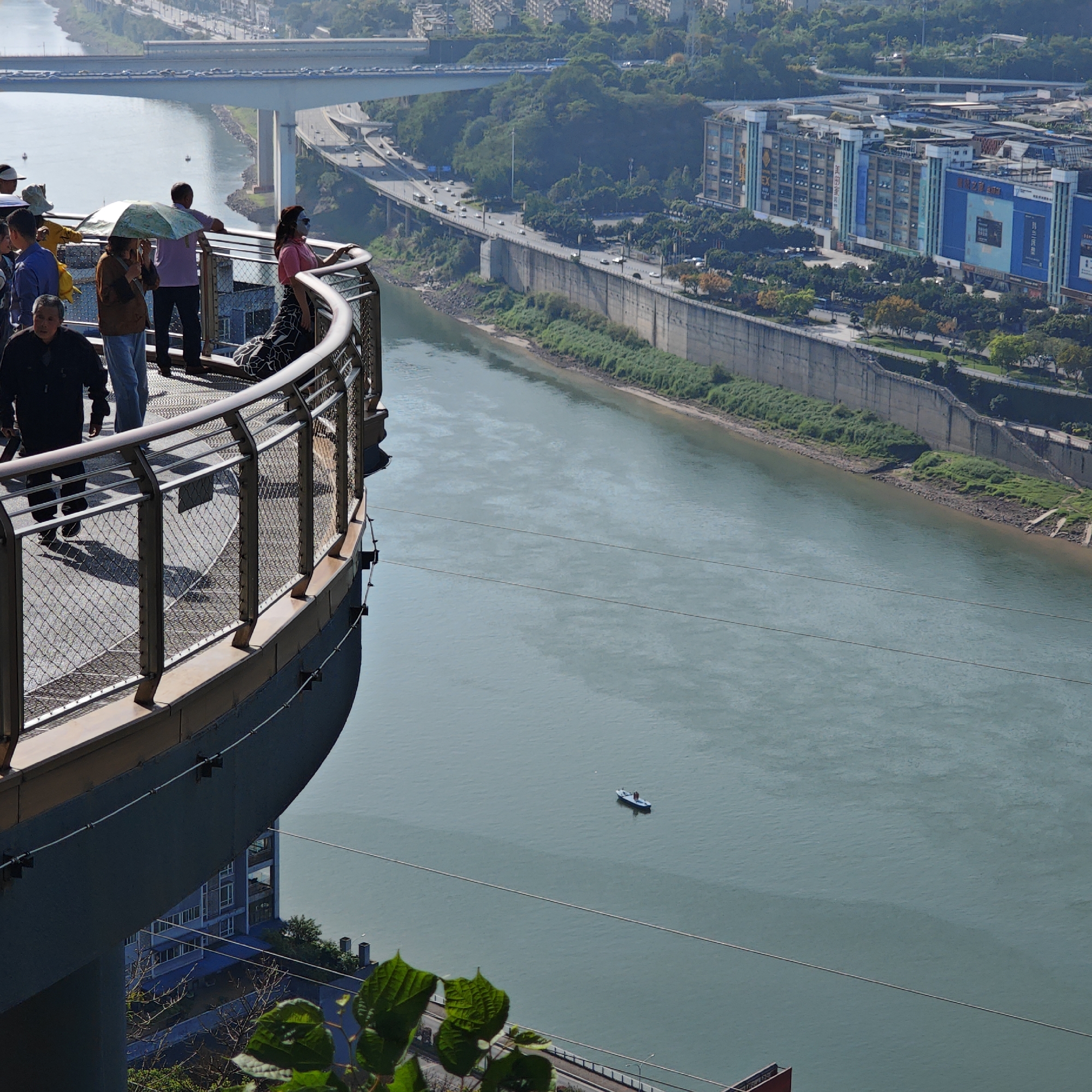 A high-angle daytime shot shows a viewing platform in the city of Yuzhong District, China. Several people are on the platform, observing the river and cityscape below. To the left, a man in a black jacket walks. A woman wearing a face mask leans on the railing. In the middle, a man holds a silver umbrella. Behind them, a man in a purple shirt looks toward the river. Beyond the railing, a wide river flows between green banks and buildings. A boat with people is on the river. In the background are buildings and bridges, with roadways visible along the riverbank.