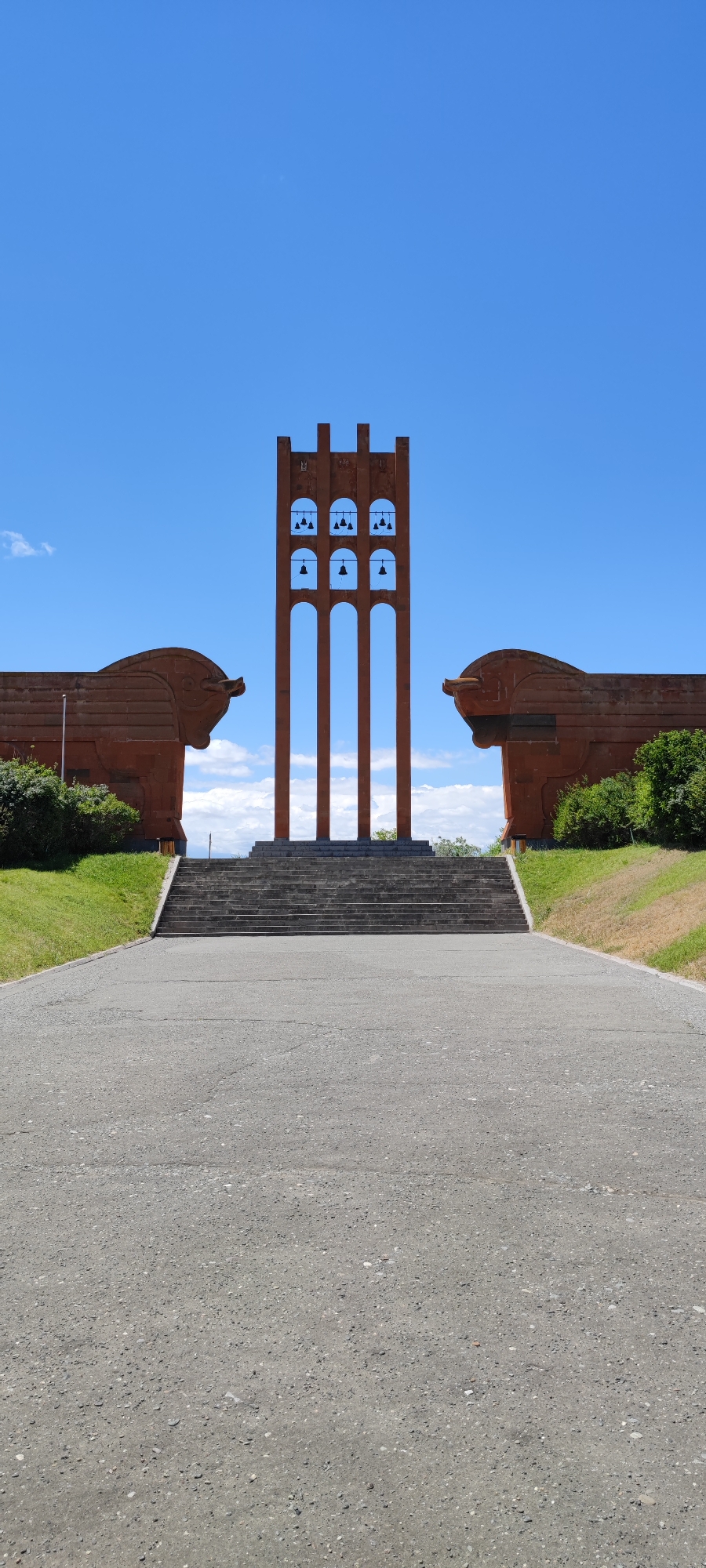 The image captures a monument located in the city of Araks, Armenia. In the foreground, a paved pathway leads towards a wide set of concrete stairs. The stairs ascend to a raised platform where a striking bell tower stands prominently. The tower is made of a reddish-brown material and features multiple arched openings, each containing bells. On either side of the bell tower, symmetrical, large, sculpted figures resembling horse heads, also made of the reddish-brown material, flank the scene. Behind the monument, a clear blue sky with scattered white clouds provides a backdrop. Green grass covers the ground around the base of the monument and the sculpted figures.