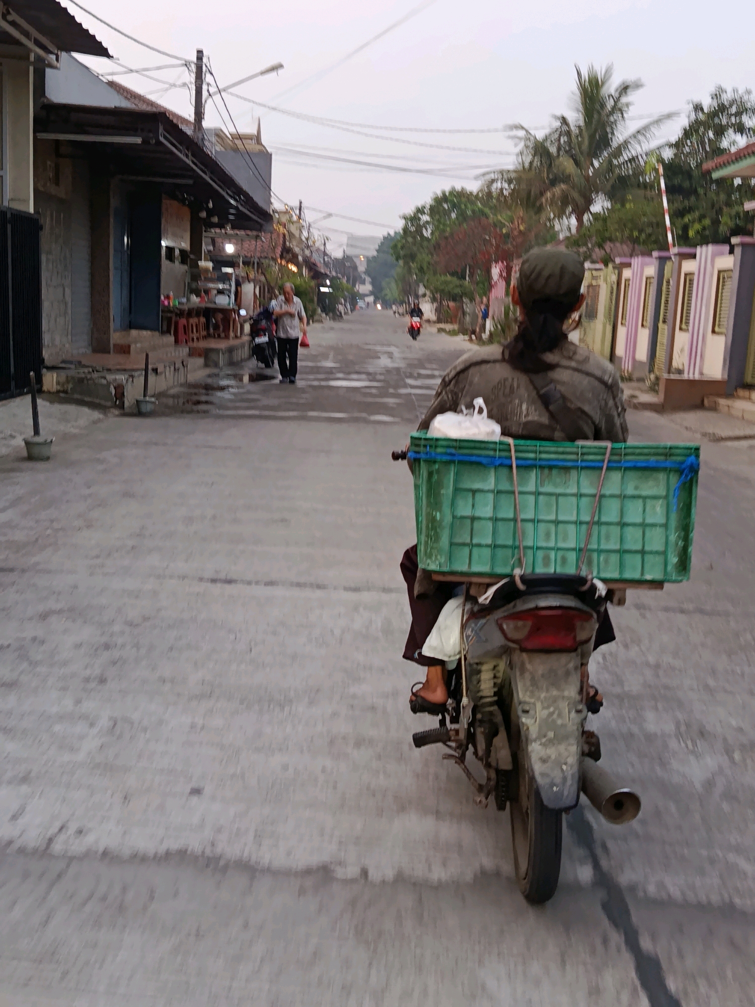 A person wearing a dark cap and jacket rides a motorcycle down a paved street in Bekasi, Indonesia. The motorcycle has a large green plastic crate attached to the back, holding a white plastic bag. The street is lined with buildings, some with awnings and others with colorful striped facades. A man in a white shirt and dark pants walks on the sidewalk in the distance. Electrical wires crisscross overhead. The sky is overcast, suggesting either early morning or late afternoon.