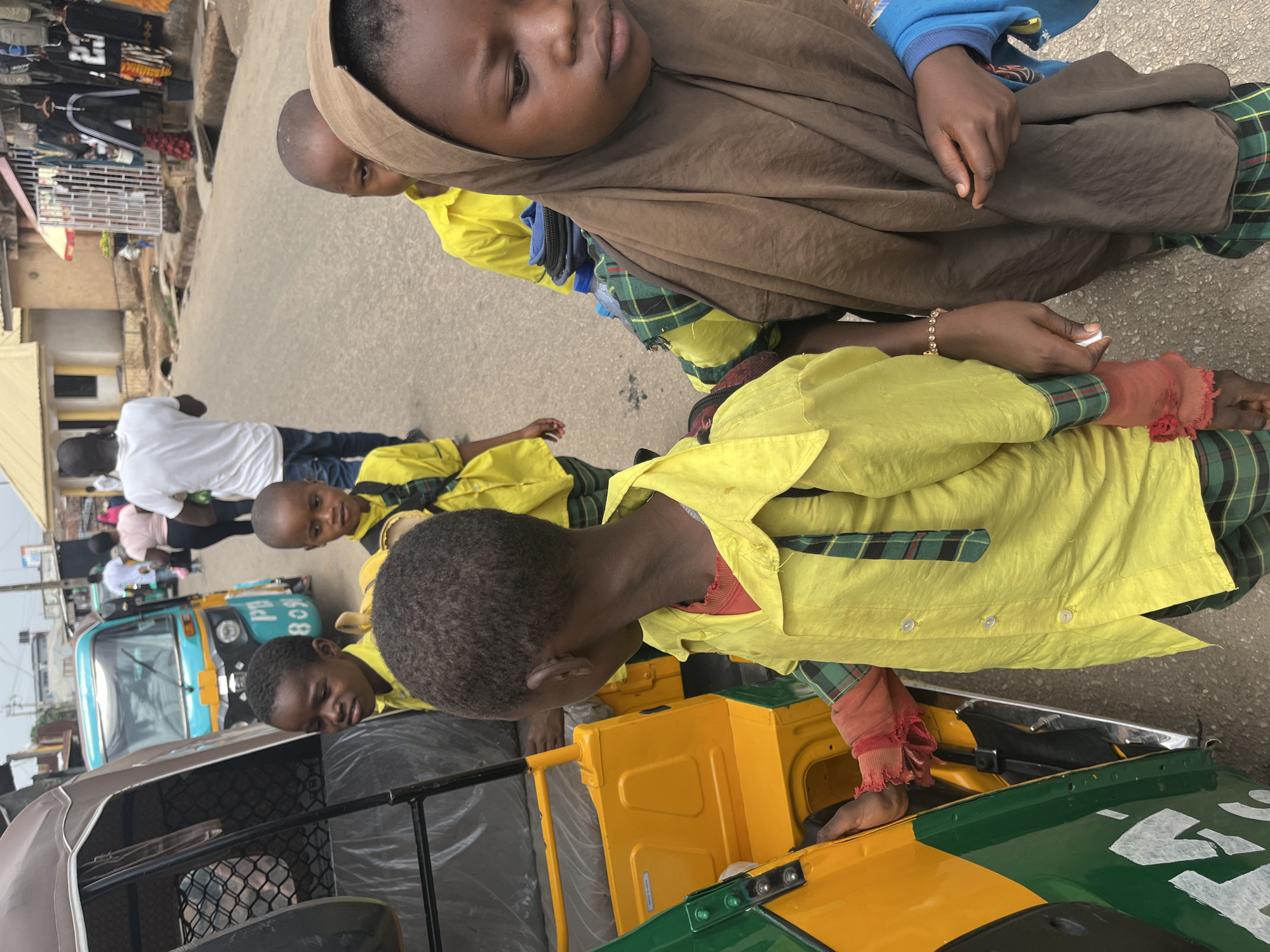 Several children are gathered on a street in Gura Top, Nigeria, near parked auto rickshaws (tuk-tuks). The children are wearing yellow shirts with green and black plaid patterns. One child wears a brown hijab. One of the children is seated inside one of the auto rickshaws, placing their bare foot on the vehicle's exterior. The scene appears to be an ordinary street in a Nigerian city, with buildings, street vendors and pedestrians visible in the background.