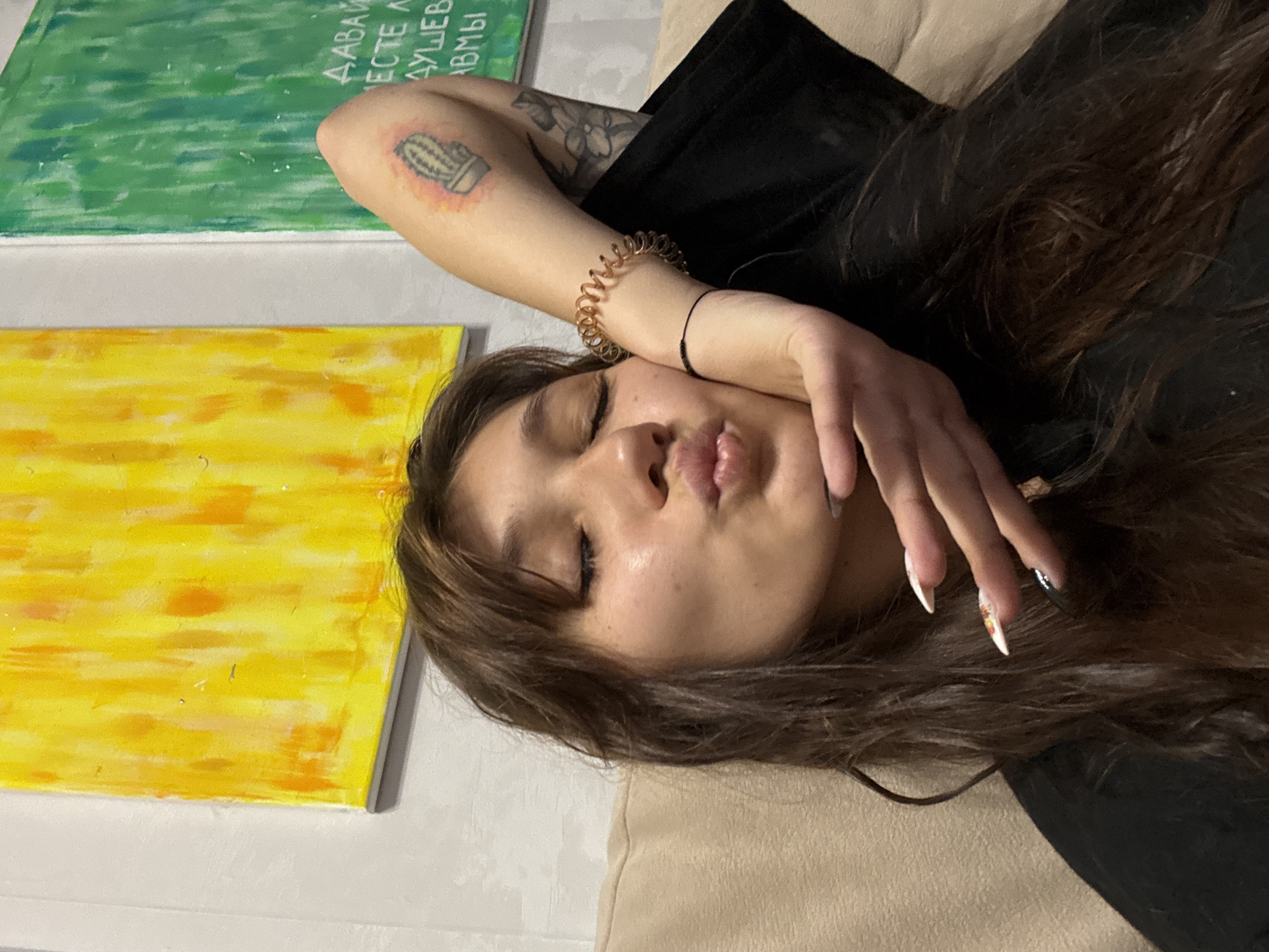 A young woman with long, dark brown hair and bangs is taking a selfie. Her eyes are closed, and she is puckering her lips. Her right hand is raised, with her fingers gently touching her chin. She has long, manicured fingernails with a white and colorful design. She is wearing a black top. On her right arm, she has a tattoo of a cactus and other floral tattoos. She is also wearing a gold, coiled bracelet on her right wrist. Behind her, there are two abstract paintings on the wall, one yellow and orange, and the other green. The scene appears to be indoors, likely in a home, given the presence of the paintings and the casual setting. The background is slightly blurred, suggesting a shallow depth of field. There is no indication of the city of Novokuznetsk, Russia, in the image.