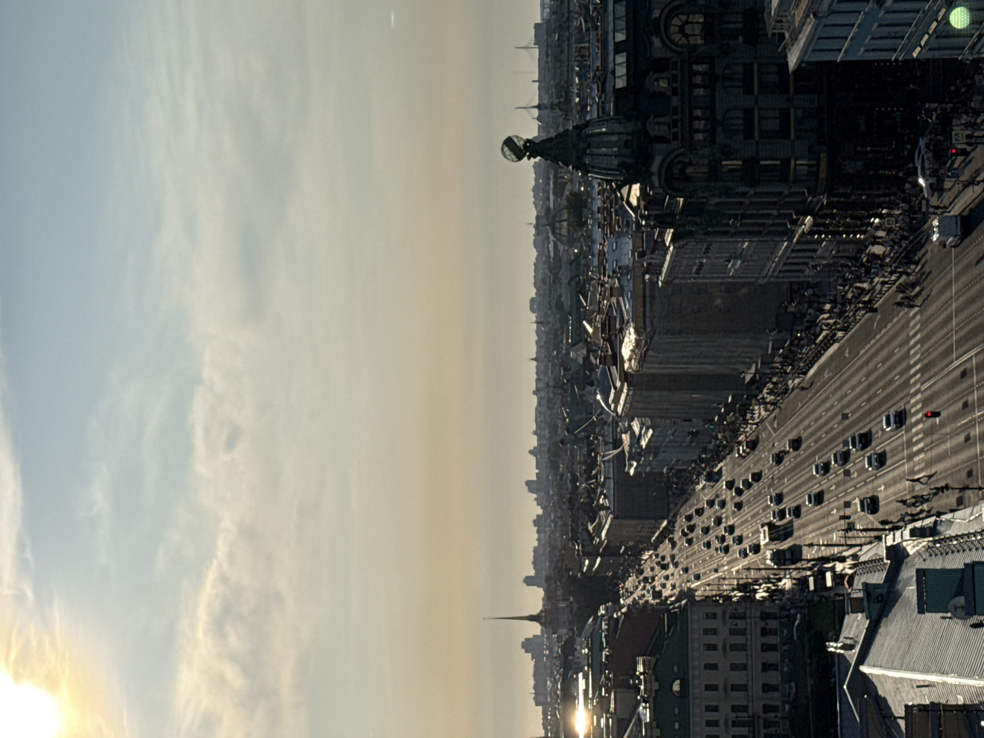 This is a high-angle shot of a wide city street in Saint Petersburg, Russia, likely Nevsky Prospekt, during daylight. The sun is low, casting long shadows and creating a warm, golden light across the scene. The sky is a pale blue with wispy clouds. The street is lined with multi-story buildings with ornate architecture, typical of the city. Traffic, consisting of cars and a bus, moves along the street, which appears to be a major thoroughfare. Pedestrians are visible on the sidewalks. In the distance, the city skyline includes various buildings and spires, with a prominent pointed spire in the center-left and a building topped with a globe on the right. The overall impression is of a bustling urban environment bathed in the soft light of either early morning or late afternoon.