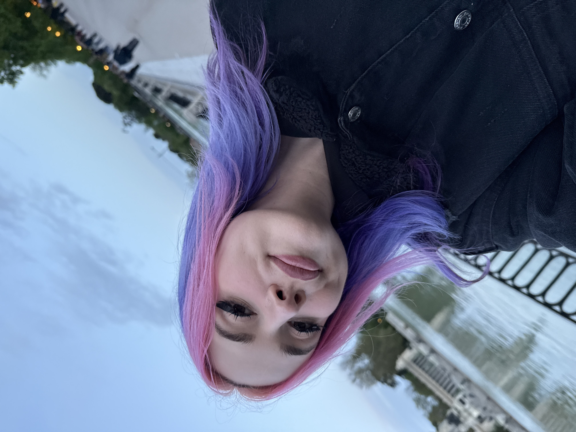 A young woman with dyed pink and purple hair is taking a selfie. She is wearing a black jacket. Behind her is the Estanque Grande (Great Pond) in El Retiro Park, Madrid, Spain. People are visible in the background along the edge of the pond, near the monument to Alfonso XII.