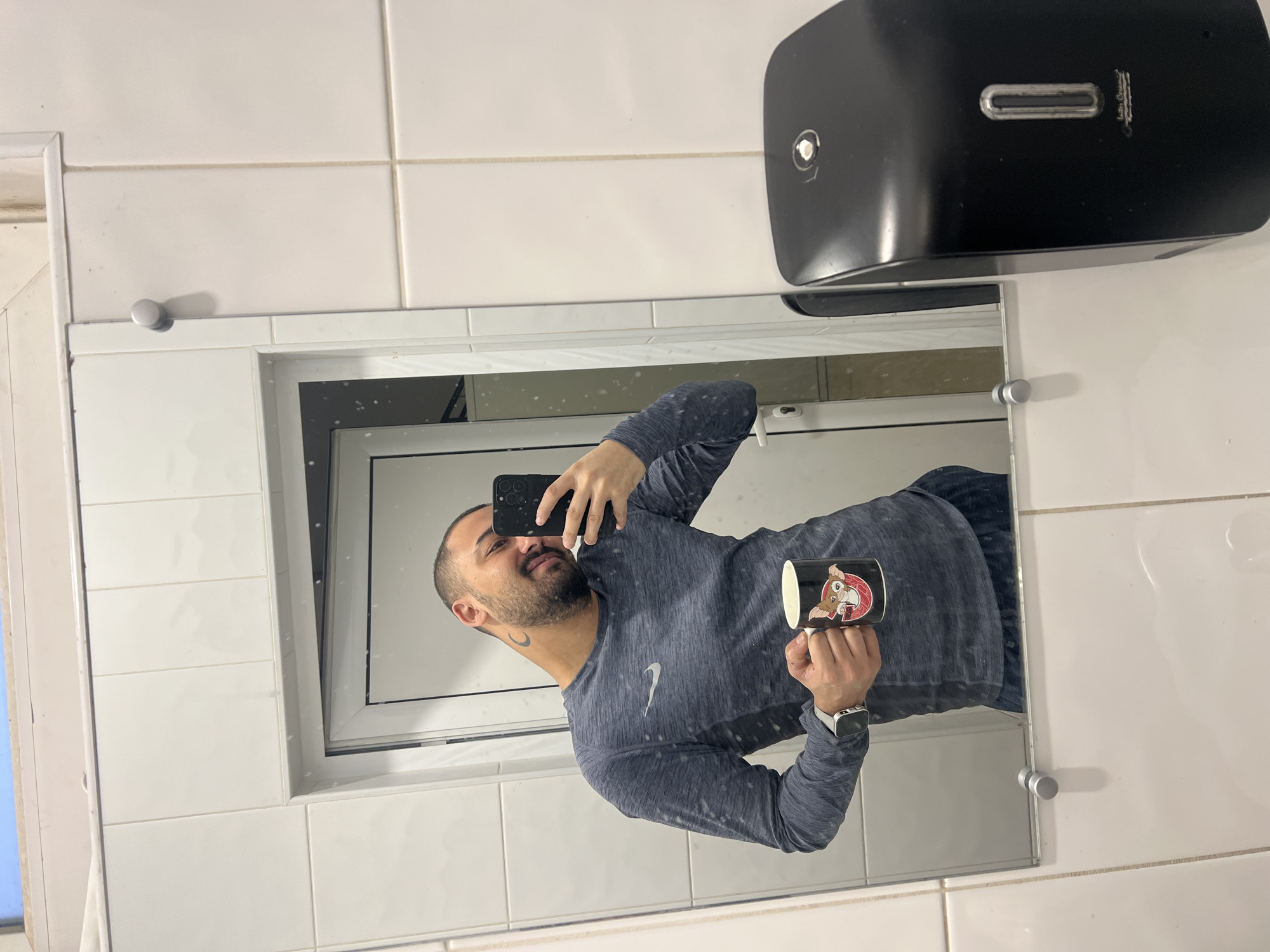 A man is taking a selfie in a bathroom mirror. He is holding a black phone in his left hand and a mug with a cartoon character on it in his right hand. He is wearing a grey long-sleeved shirt with a white swoosh logo and a white smartwatch. The bathroom has white tiled walls, and a black soap dispenser is mounted on the wall above the mirror. A white window frame is visible in the reflection. The location in the city of Sevlievo, Bulgaria is not discernible from the image.
