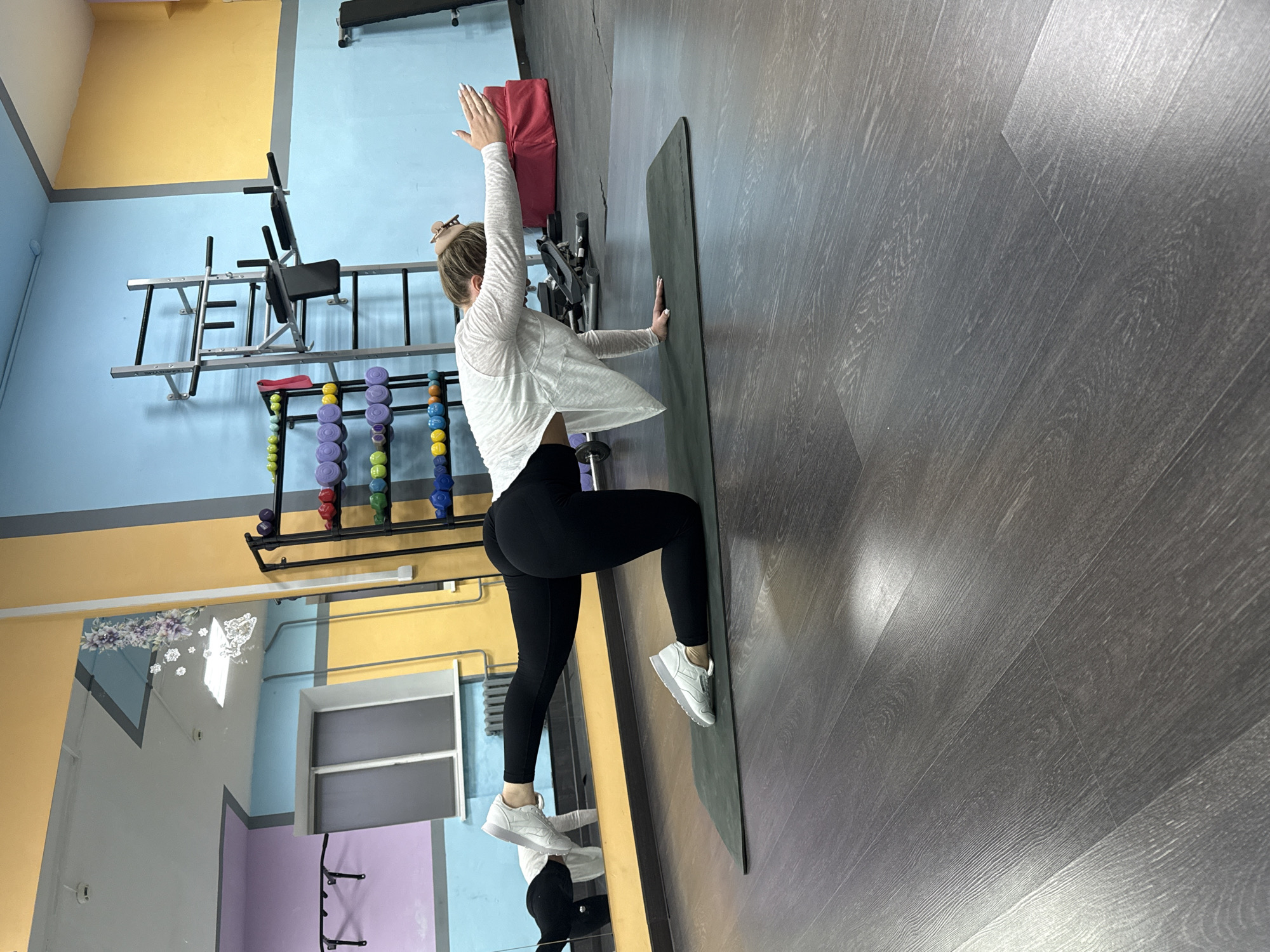 A woman is stretching in a gym located in Chaikovsky, Russia. She is standing on a dark grey yoga mat in a lunge position, with her left arm raised and right hand against the wall, stretching her torso. She is wearing black leggings, white sneakers, and a light-colored cardigan. The gym has blue, yellow, and grey walls. Exercise equipment is visible in the background, including a pull-up bar, weight racks with dumbbells, and a mirror.