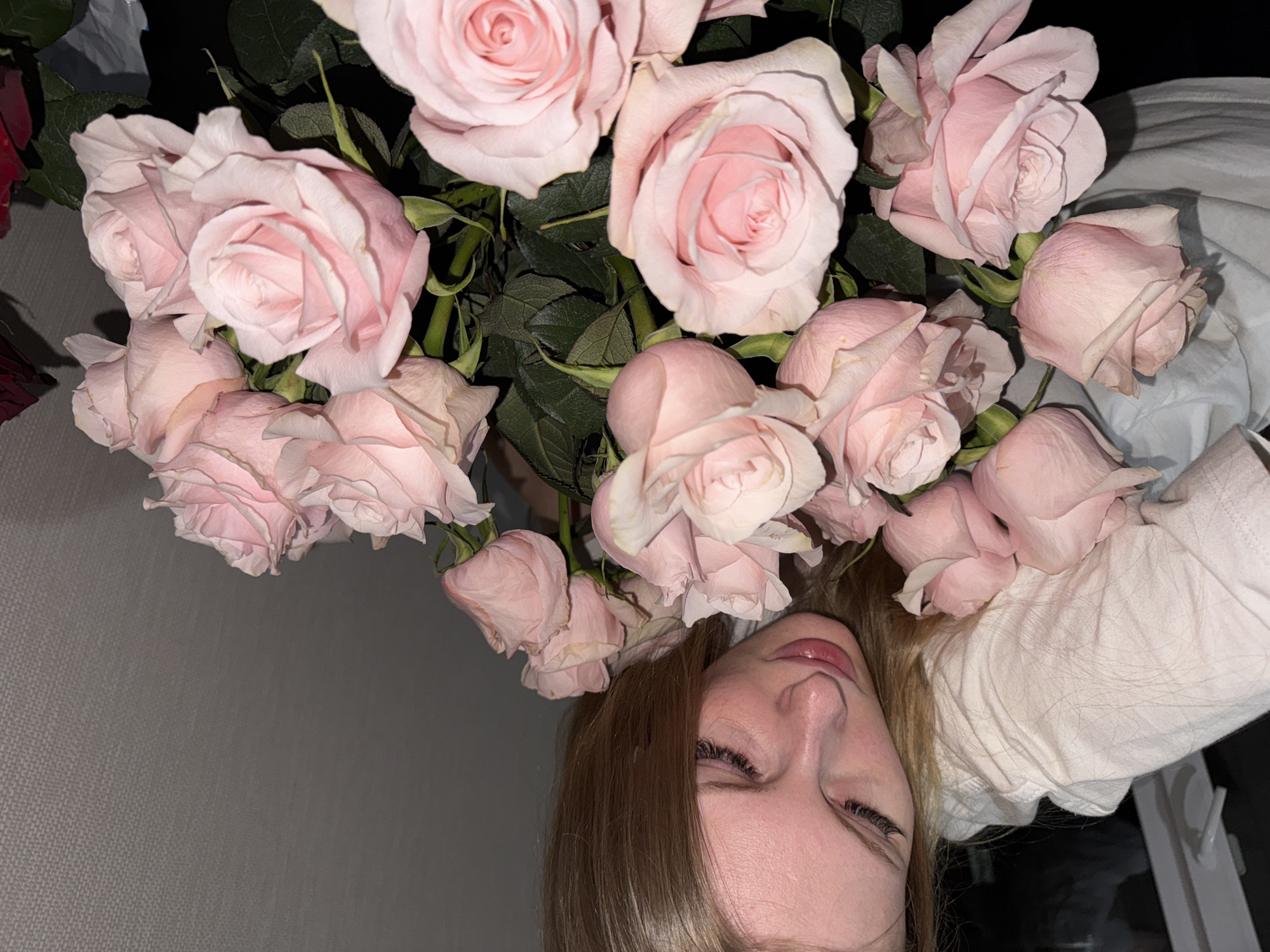 A young woman with long blonde hair is seen in a close-up shot, looking down at a bouquet of light pink roses. She is wearing a white hoodie. The background is a textured grey wall. The scene is indoors, likely in an apartment, with no specific Perm landmarks visible. The lighting is dim, suggesting it might be evening or nighttime.