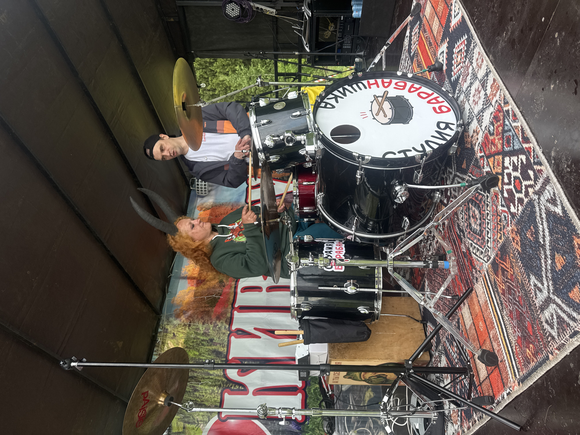 An outdoor stage in Большая Кивара, Russia, features two individuals interacting with a black drum kit. A woman with reddish-orange curly hair and dark horns, wearing a green hoodie and blue pants, is seated at the drum kit, holding drumsticks. To her right, a man with dark hair, wearing a black backward baseball cap, a white t-shirt, and a dark jacket with orange and white stripes, stands behind the kit, also holding drumsticks.
The drum kit consists of a bass drum with a white head featuring a "СТУДИЯ БАРАБАНОВ" (STUDIO OF DRUMS) logo and a drum illustration, a rack tom with a "Студия Барабанов" sticker, a floor tom, and a snare drum. Various cymbals, including a Paiste crash, a hi-hat, and a ride, are mounted on stands. A black fabric stick bag hangs from a stand on the left. A light brown wooden cajón with a frog graphic and "А ТЫ СЕБЯ" text is positioned beside the drum kit on the left.
The stage floor is dark, overlaid with a multi-colored geometric pattern rug beneath the drum kit. The backdrop is a large banner displaying the red and white word "КИВАР" with a partial Pepsi logo, set against a forest scene with green and autumn-colored trees. The stage has a dark, covered ceiling structure. Additional stage equipment, including a lighting fixture and cables, is visible on the right. Green trees are discernible beyond the stage structure, indicating an outdoor setting. Two blue beverage cans rest on the stage floor on the far right.