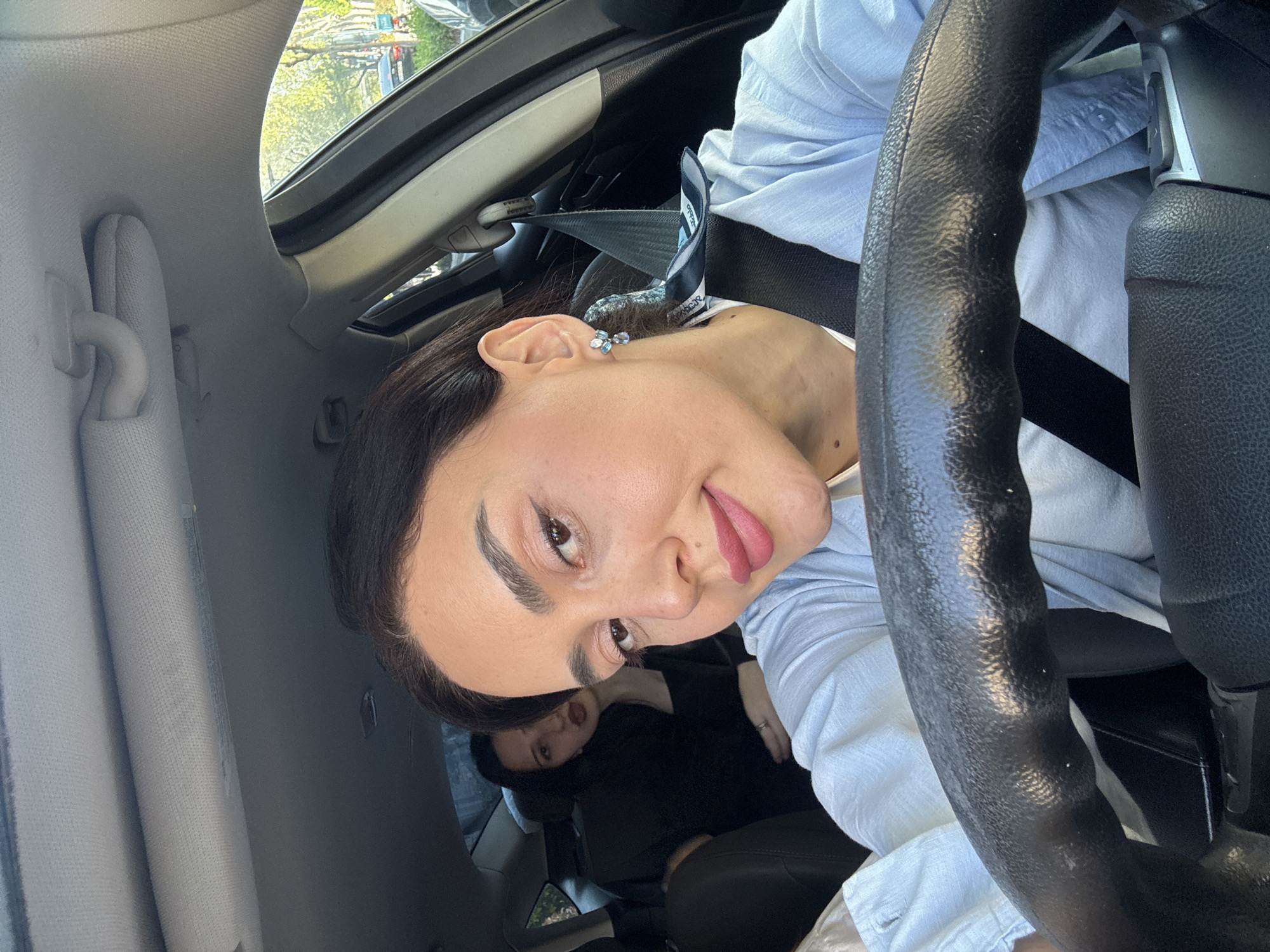 A playful selfie taken inside a car shows a woman in the driver's seat looking directly at the camera with her head tilted, a slight smile, and her tongue subtly out. She has dark hair, pronounced eyebrows, and is wearing light blue button-up shirt and blue and silver floral-shaped earrings. A black seatbelt crosses her chest. The black textured steering wheel is prominent in the right foreground.
In the background, another person with dark hair is visible in the back seat, looking forward. The car's interior features a light gray fabric headliner and sun visor, with a partial view of an open sunroof revealing green trees and what appears to be a sunny day outside. The car's seats are black. The setting is a car interior during daytime, in Tbilisi, Georgia.