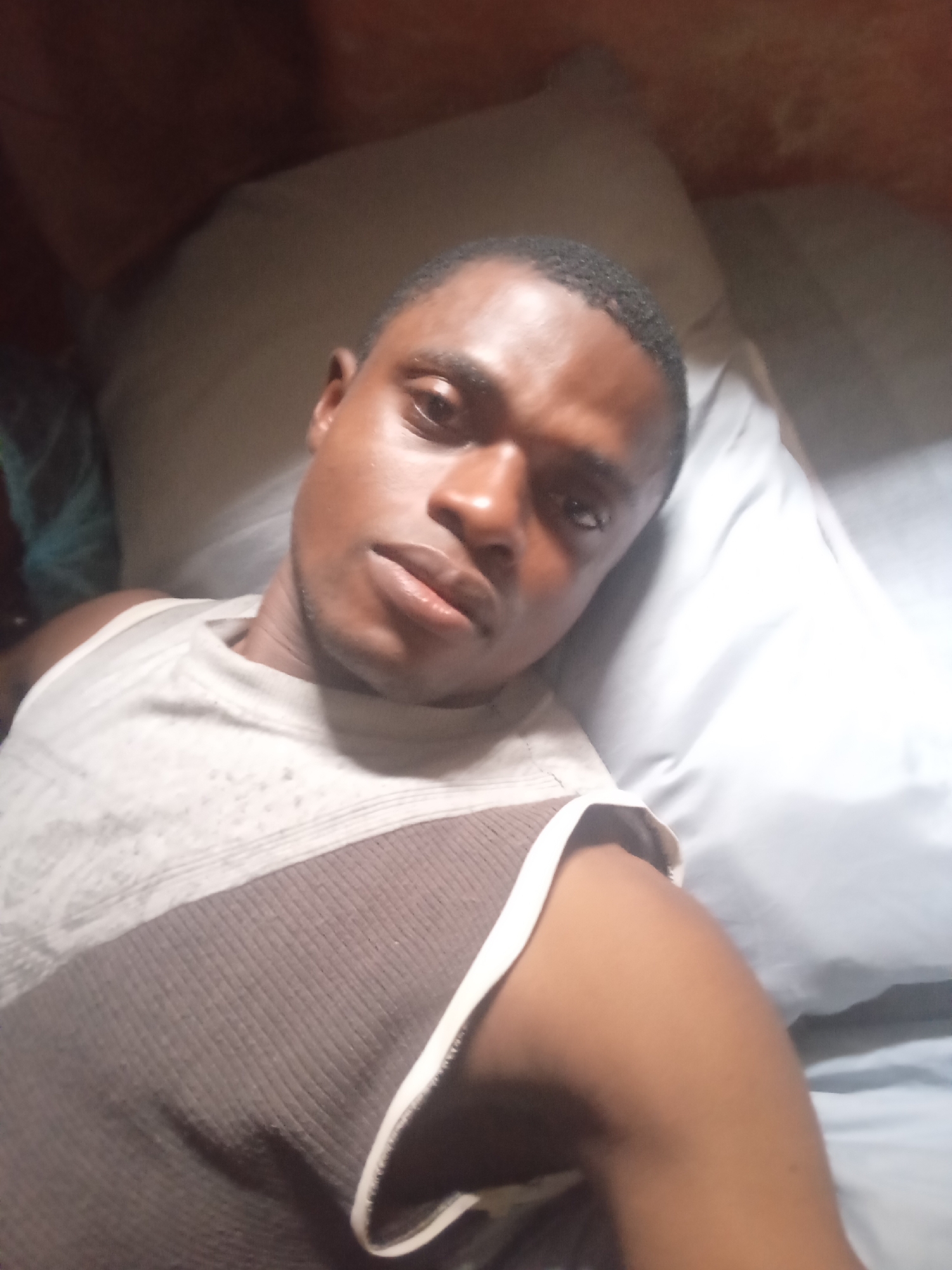 The image is a close-up selfie of a dark-skinned man lying on his back. He is wearing a sleeveless shirt with a brown and white striped pattern. The background appears to be a dimly lit room with pillows and what looks like a wooden ceiling. The man's head is tilted towards the camera, and his eyes are open. He has a neutral expression on his face. The lighting is soft, casting shadows on his face and body. The image quality is moderate, with some graininess. The location in the city of Ashipa, Nigeria, is not discernible from the image itself.