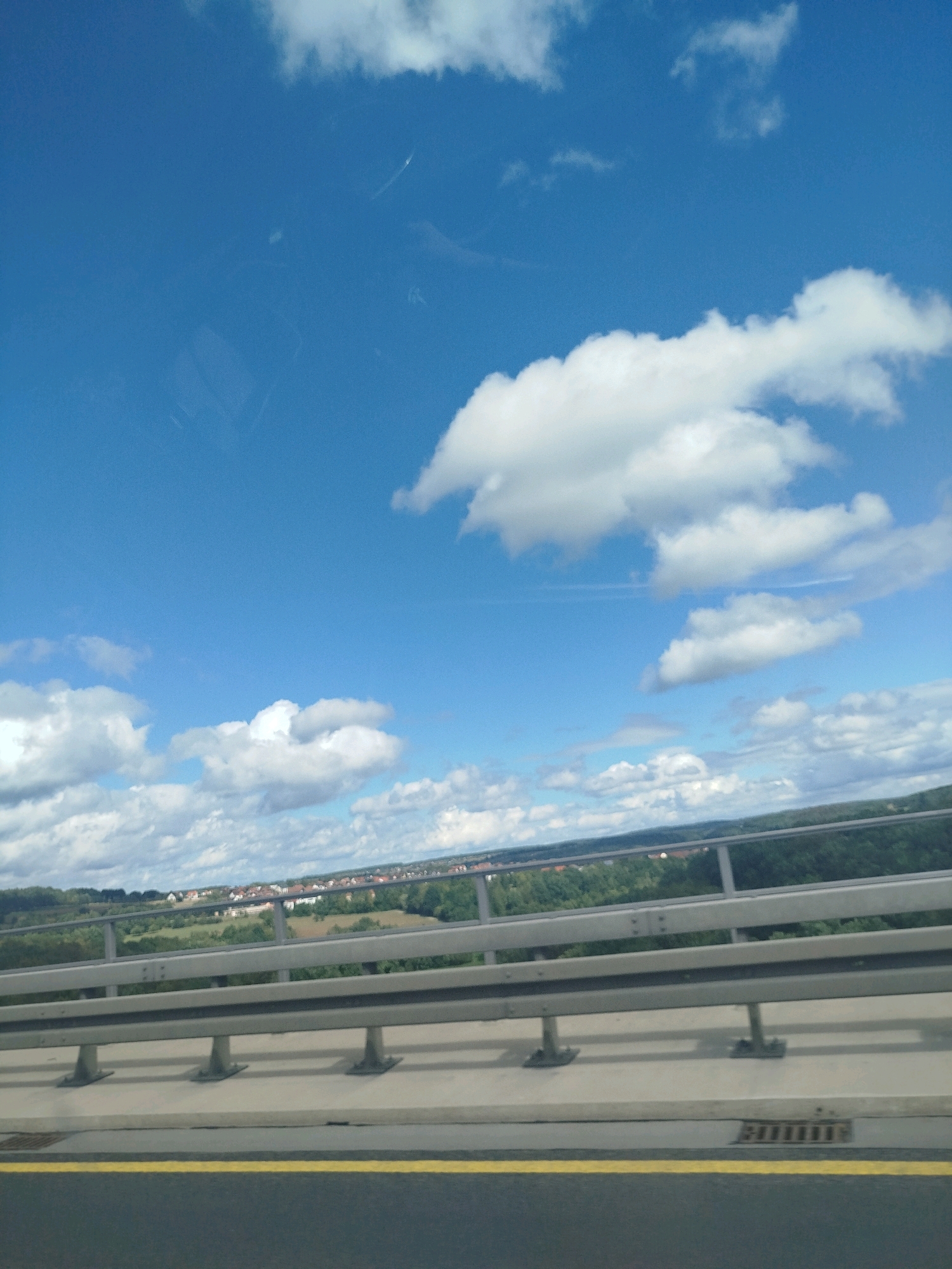 The image is a photograph taken from a moving vehicle on a highway overpass, looking out at a landscape under a bright blue sky with scattered white clouds. In the foreground, a concrete barrier with a silver metal guardrail, supported by metal posts, is visible. A yellow line runs along the edge of the dark asphalt road. In the mid-ground, a suburban area with red-roofed houses is nestled amongst green trees and fields. The sky occupies the upper portion of the image, with a faint contrail from an aircraft visible in the upper left quadrant. The scene suggests a view of the outskirts of Oberthulba, Germany, likely taken during daylight hours with good weather conditions.
