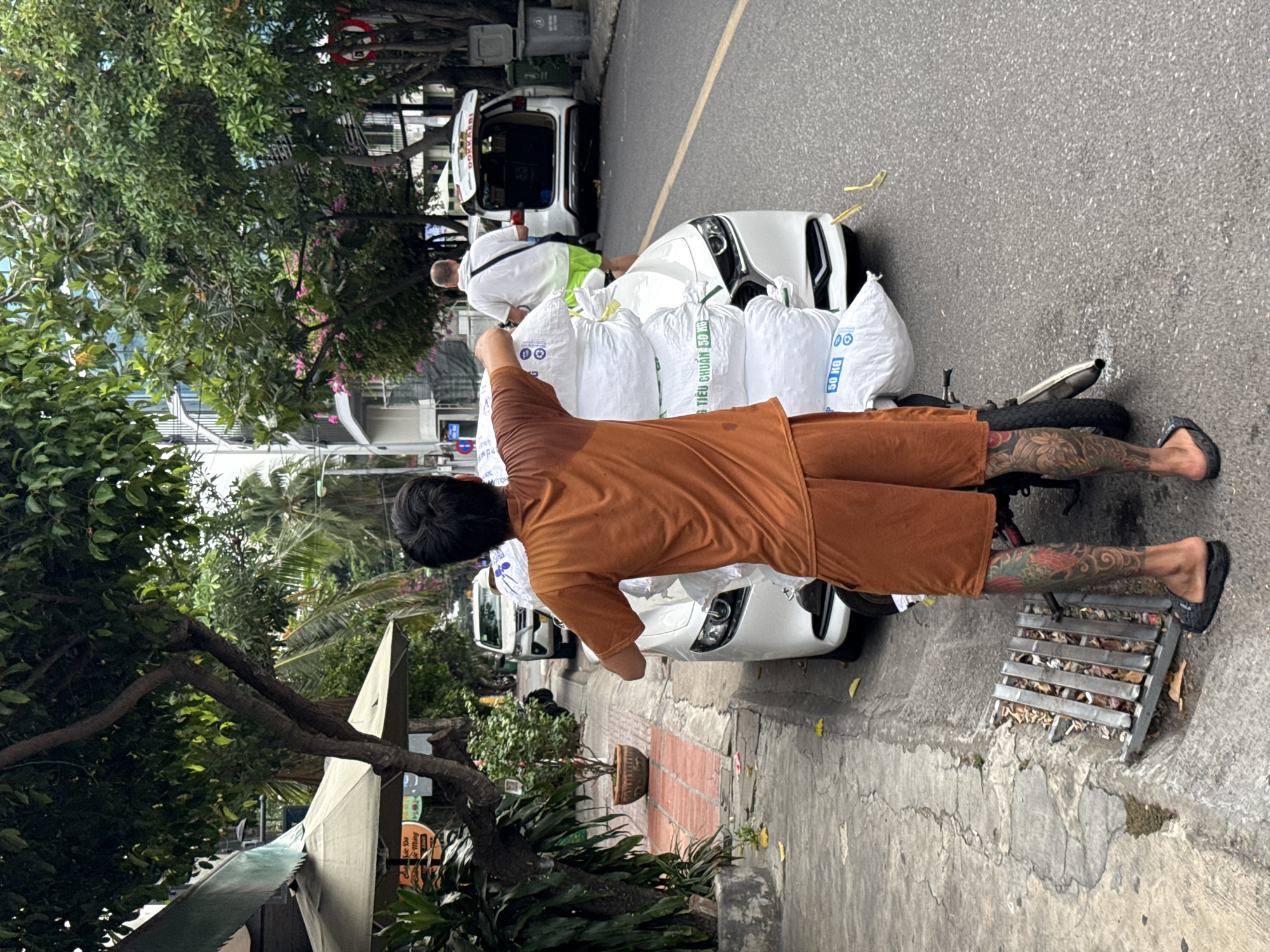 In Nha Trang, Vietnam, on a city street lined with trees and buildings, a man with extensive leg tattoos is seen from behind, wearing an orange t-shirt and shorts. He is standing next to a white car, on which are stacked several large white sacks. The sacks are labeled with Vietnamese text and "50 KG". Another man in a white shirt and green shorts is walking away from the camera toward a white SUV with its trunk open. The street has a concrete sidewalk with a metal grate.