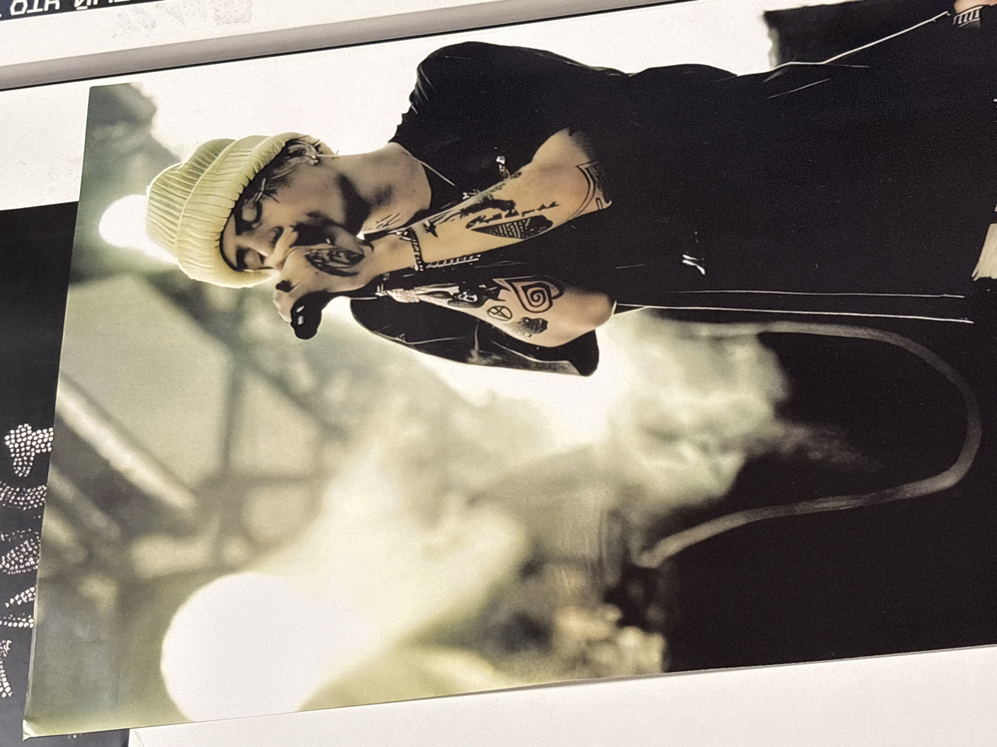 The image depicts a man with numerous tattoos on his arms and face, wearing a knit cap, holding a microphone and appearing to be performing. The background is blurred, suggesting a stage or performance venue. The scene's location in Прикамский, Russia is not evident from the image.