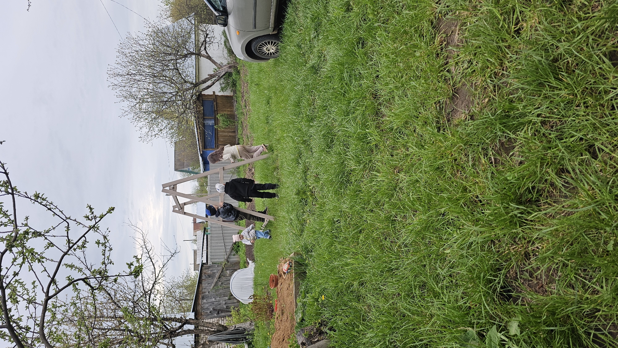 The image depicts a group of four people in a grassy yard. An adult female is standing at a swing set. There's an adult male standing nearby. In front of the swing set, there is a person wearing a blue hat and a child with a pink hat. In the background are rural structures and a partially visible car. The scene appears to be a backyard in an area with simple buildings, possibly in Neftekamsk, Russia, given the prompt's specification. A bare tree is in the foreground and a similar tree with budding leaves in the background. There is also a garden section with plastic covering next to a wooden building.