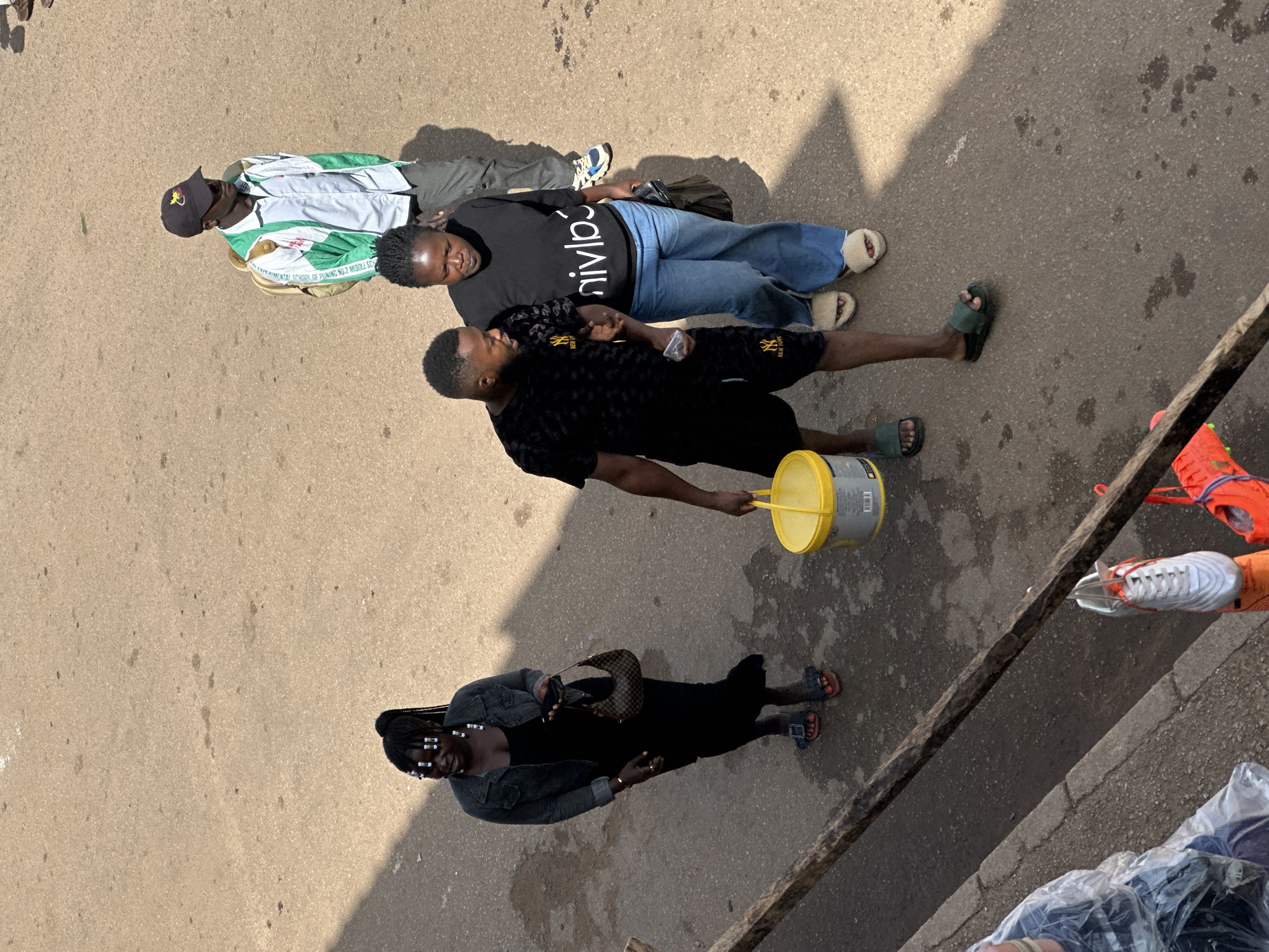 In Bukuru, Nigeria, on a dirt road, three adults and one child walk. The child, in the foreground on the left, is wearing a clear plastic bag over their head and torso. To the right of the child, a woman with braided hair and white beads walks ahead, carrying a brown purse. She is wearing a black dress and a denim jacket. Following her is a man in a black t-shirt with a repeating yellow logo and black shorts. He is carrying a yellow bucket in his left hand and appears to be holding the woman's hand with his right. Behind them, a woman in a black t-shirt with white lettering and blue jeans walks, wearing white fluffy slippers. Further back, a man wearing a green and white jacket, a black baseball cap, and gray pants walks with a light brown backpack. The ground is dusty and has some darker patches. In the lower right, an orange shoe is visible. In the lower left, a white shoe is hanging from a wooden beam.