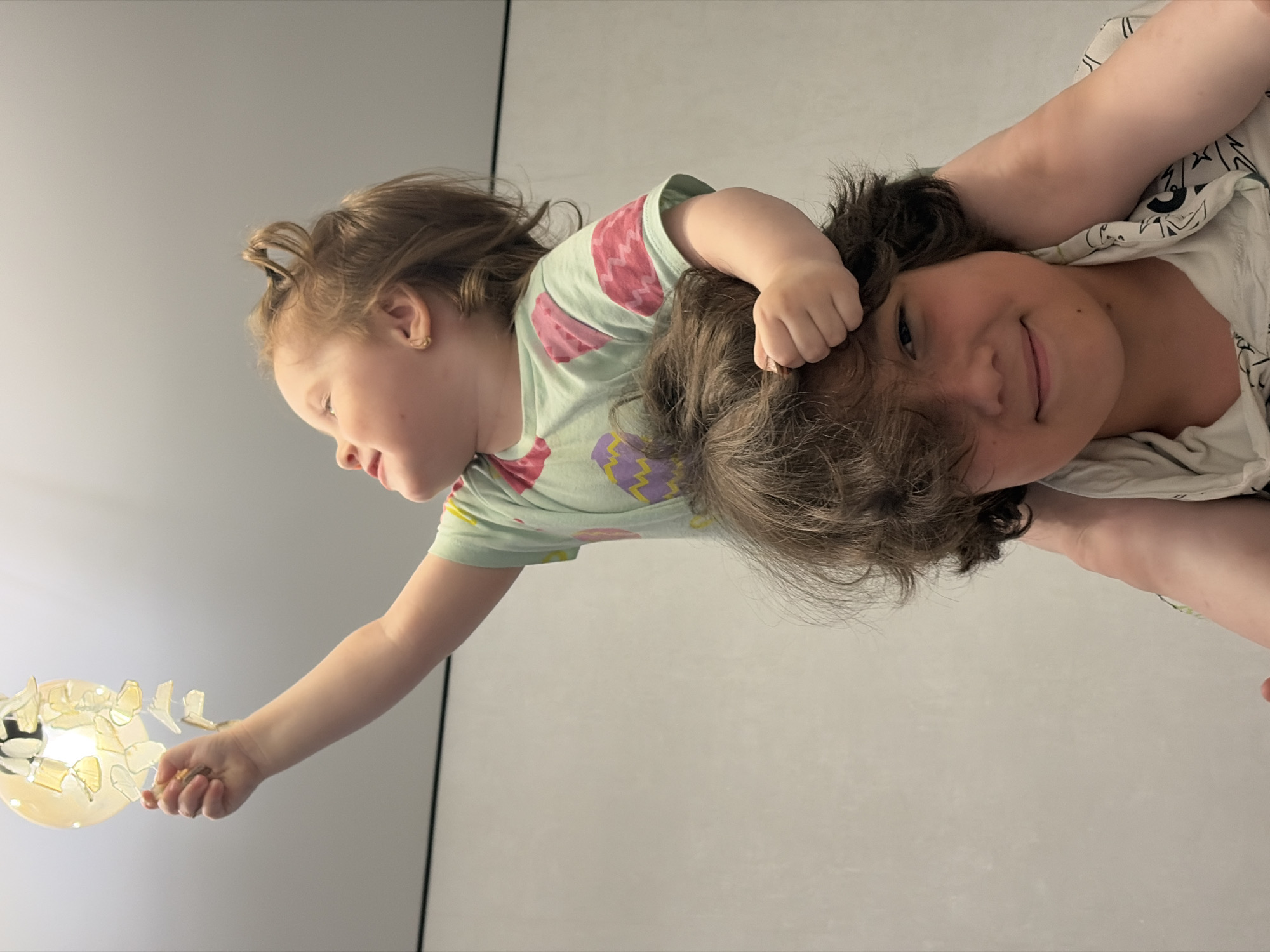 A low-angle, medium shot captures two children in an indoor setting. The child on top, a young girl with reddish-brown hair, is perched on the shoulders of the child below, a boy with dark, messy hair. The girl is reaching up with her right hand towards a decorative light fixture, her arm extended. She is wearing a light green t-shirt with a repeating pattern of pink and purple Easter eggs. The boy's face is visible as he smiles at the camera, with his arm bent and hand resting on the back of the girl's head. The background is a plain, light-colored wall. There are no visible landmarks to suggest the location in Chaikovsky, Russia.
