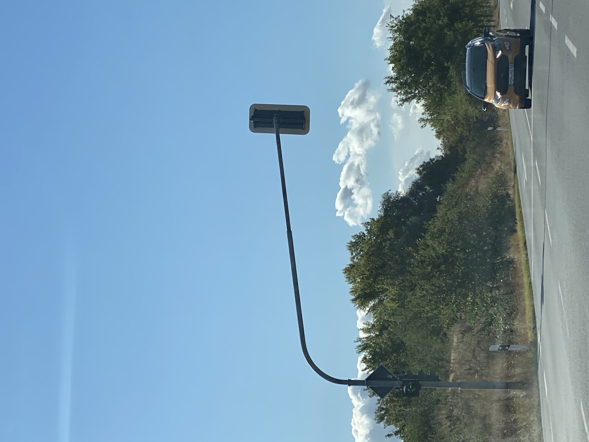 This is a full-frame, eye-level, outdoor, daytime photograph taken in Nordhausen, Germany. The image features a road with a single lane in the foreground, bordered by a grassy verge and trees. A golden-orange SUV, with license plate "NDM HS 58," is driving away from the viewer on the road. To the left, a traffic light is visible, showing a green signal. A pole with a diamond-shaped sign stands next to the traffic light. Above the road, a long traffic signal arm extends from left to right, with a single traffic light unit suspended at its end. The background is dominated by a clear blue sky with scattered white clouds, and dense green trees line the right side of the road.