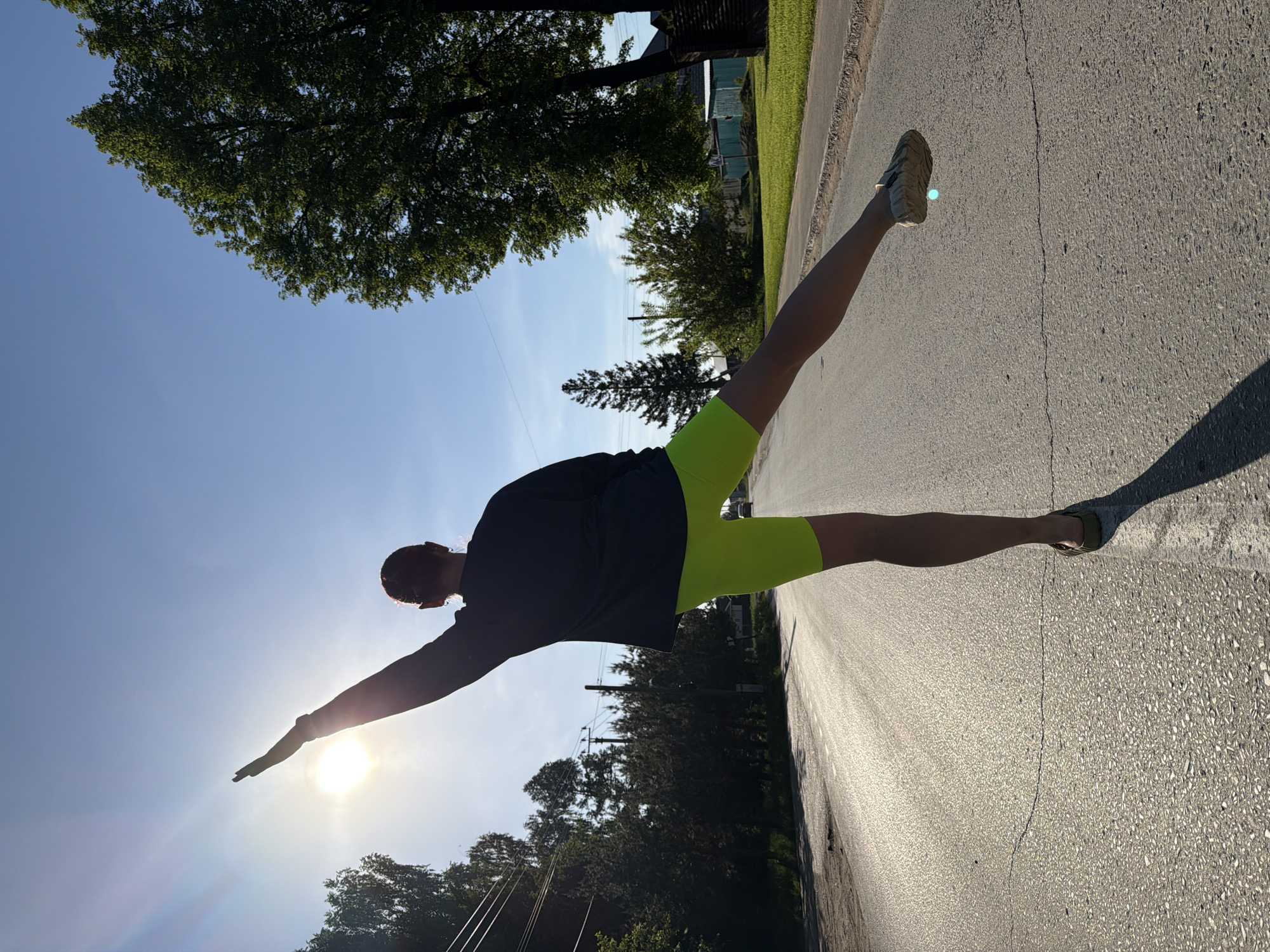 A person wearing a dark top and neon green shorts is performing a split on a paved road. The right arm is extended toward the sun. The background consists of trees, utility poles, and buildings. The location is the city of Chaikovsky, Russia.
