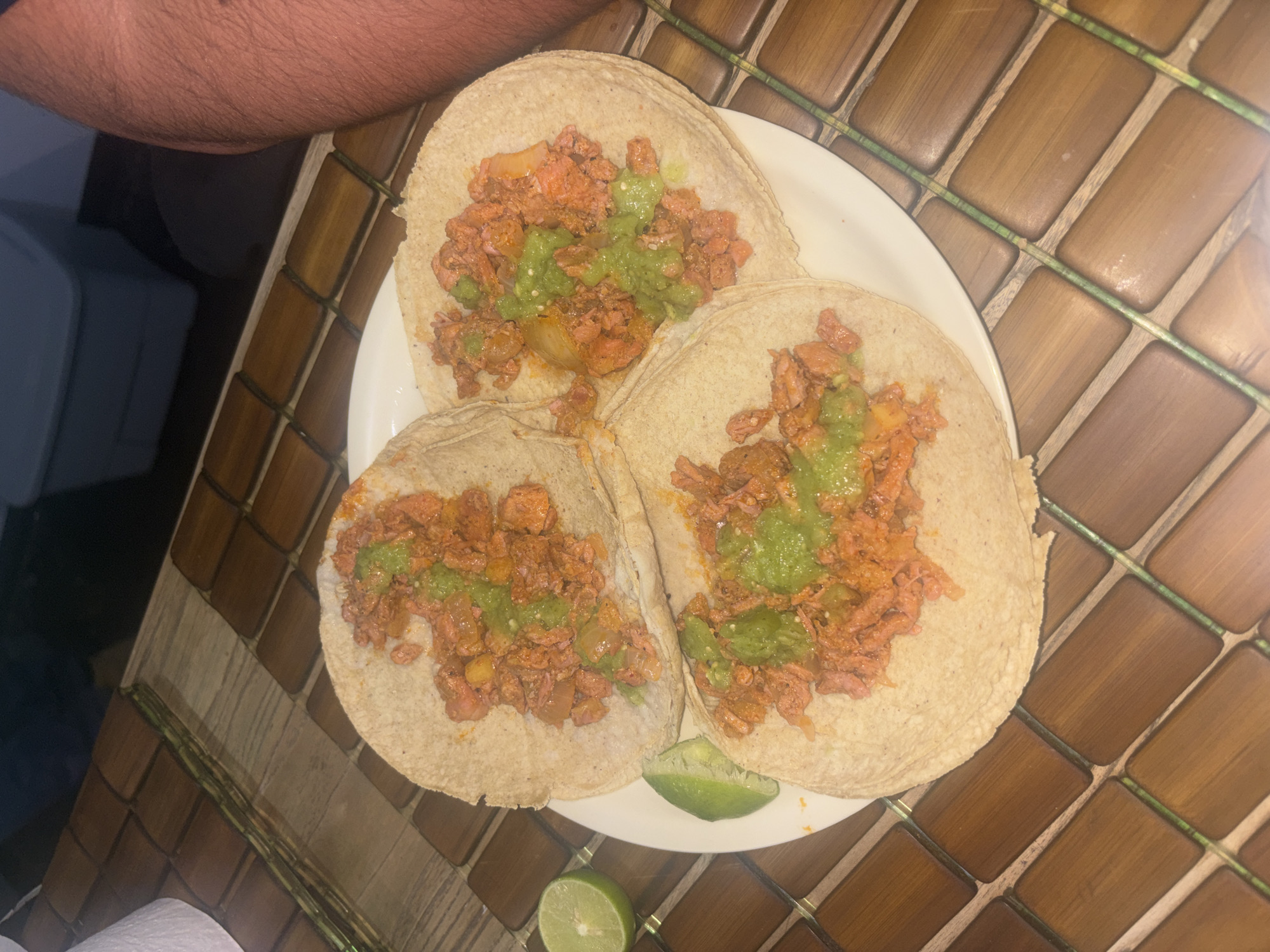 The image shows a plate of three tacos with orange-colored meat, green sauce, and two lime wedges on a wooden table. The location is Buenavista, Mexico. A person's arm is visible in the upper-left corner of the image.