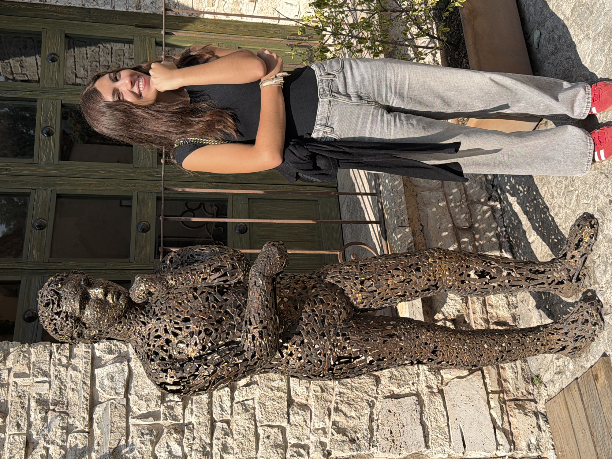 The image shows a woman lying next to a metal sculpture of a person, likely in Domingo Arenas, Mexico. The woman is smiling at the camera, wearing a black top, grey jeans, and red shoes. She has long brown hair and is wearing bracelets on her right wrist. The sculpture is lying on a stone wall, next to the woman. The background includes a green wooden door with dark metal accents, some plants, and a stone patio. The lighting suggests a sunny day.