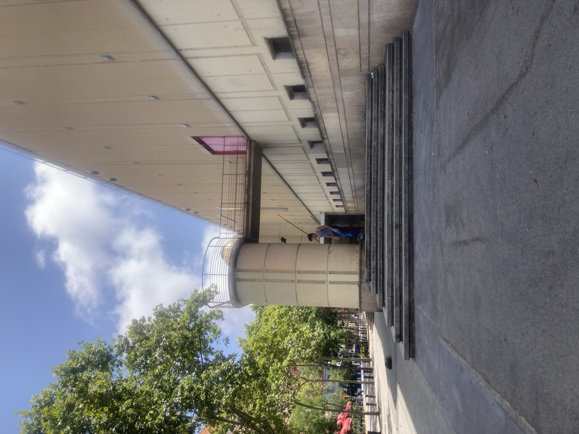 A person on a skateboard is descending a wide, shallow set of concrete stairs in front of a large, light-colored building with a red door and a walkway leading to a circular tower. The building's facade has a textured concrete finish with rectangular recesses. Lush green trees line the street in the background, where there is also a yellow vehicle and some red awnings. The sky is blue with scattered white clouds, indicating it is daytime. The overall setting suggests an urban environment in Barcelona, Spain.