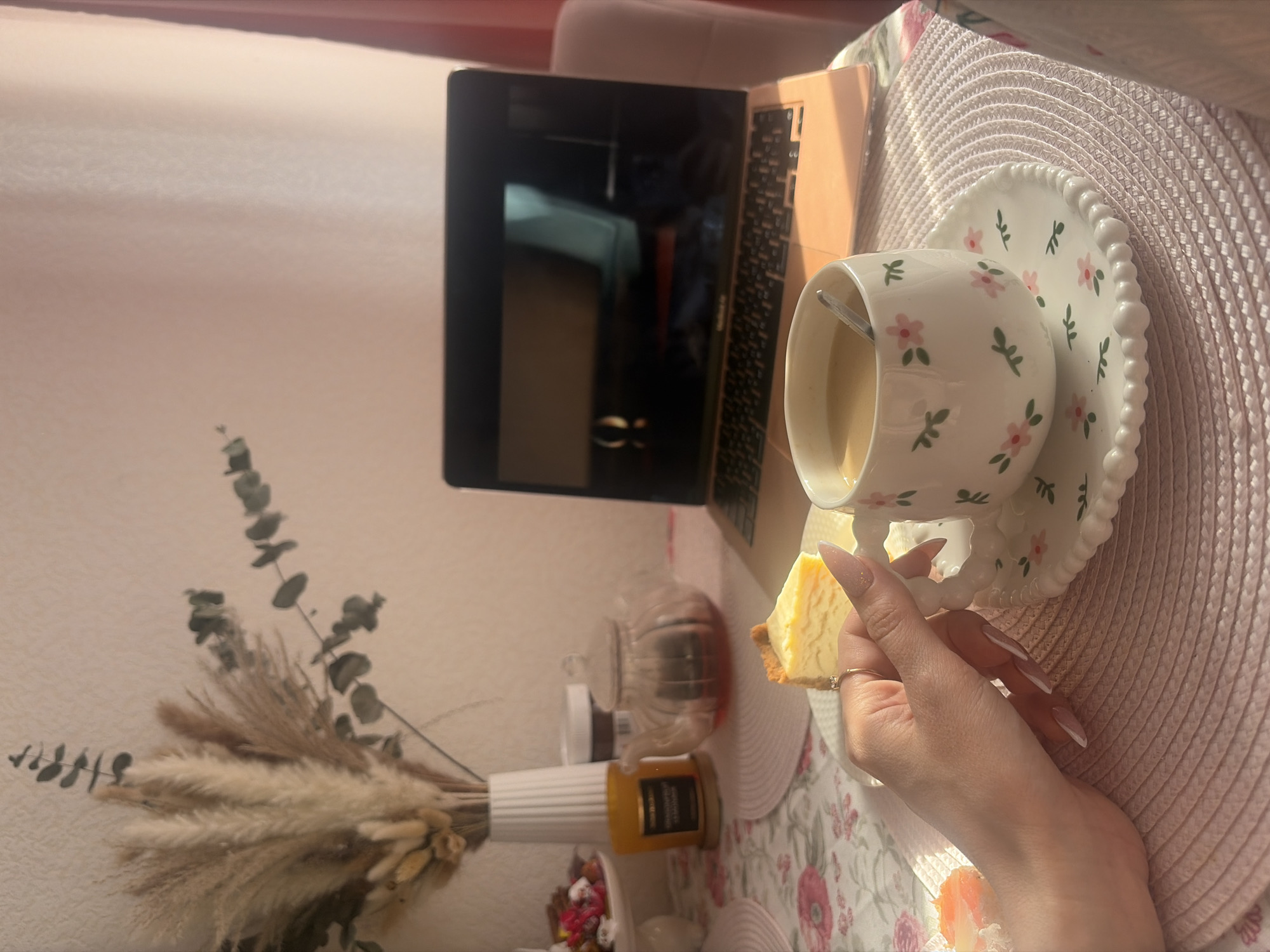 A brightly lit interior scene features a laptop, a floral teacup with a saucer, and a slice of cheesecake held by a hand with long, white-tipped nails. The objects are arranged on a floral-patterned tablecloth atop a pink woven placemat.
Behind the laptop are jars and a white vase holding dried floral arrangements, including pampas grass and eucalyptus. The wall behind the objects is off-white.
The scene is likely set indoors, with a focus on personal items suggestive of a leisure or work-from-home setting. There is no explicit indication of Saint Petersburg, Russia in the visual elements. The precise location is not discernible from the image.