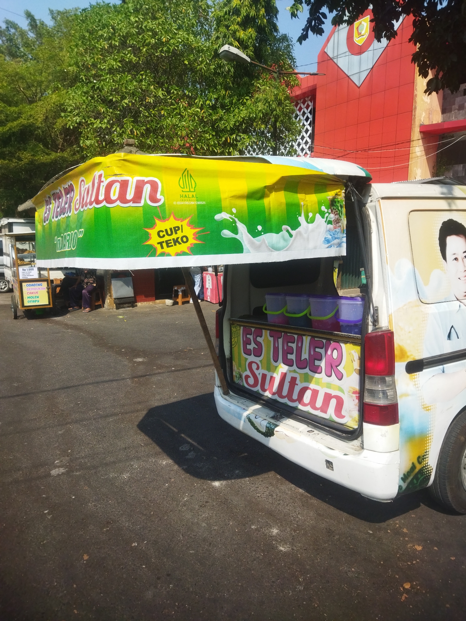 The image depicts a mobile food cart selling "Es Teler Sultan" in the city of Wonogiri, Indonesia. The cart is a white van converted into a food vendor. A large banner on the side of the van reads "ES TELUR SULTAN" in large, colorful letters. Above it, a green banner displays the words "ES TELUR SULTAN" and "HALAL" along with a certification number. The background shows a red building with a crest emblem, trees, and a street. On the cart, there are several plastic containers filled with various ingredients for the dessert. The van also features a portrait of a man on its side. The scene appears to be a sunny day, with shadows cast on the asphalt road.