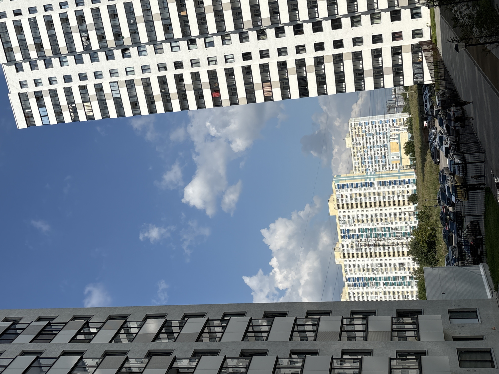 The image depicts a view of a residential area in Novosibirsk, Russia, on a clear day with scattered white clouds. Two tall apartment buildings dominate the foreground, one on the left and one on the right, framing a central vista. The building on the left is a light gray facade with many windows and balconies. The building on the right is a white facade with a grid of windows and balconies, featuring some red and pink items hanging from balconies on the upper floors. In the middle ground, a long, white apartment building with blue and yellow accents stretches horizontally, with a power line tower visible in front of it. Below this building, a field of grass and trees is present, leading to a parking lot filled with numerous cars. A black metal fence borders the parking area. In the foreground, a paved walkway leads towards the apartment buildings, with a few streetlights visible. Two figures are walking on the walkway, and another person is standing near the fence. The sky is a vibrant blue.