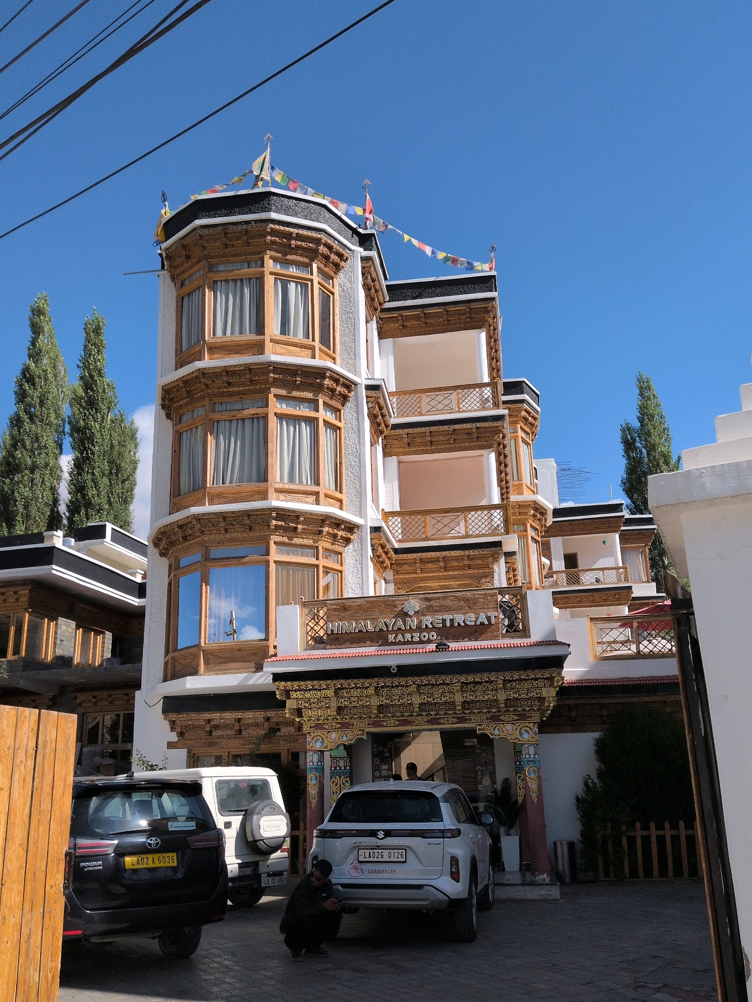 The image depicts the "HIMALAYAN RETREAT KARZOO" hotel located in Leh, India. The building features traditional Ladakhi architecture with ornately carved wooden details, multiple levels with balconies, and bay windows. Prayer flags are strung across the top of the building. In front of the hotel, a black Toyota Innova and a white Suzuki Grand Vitara are parked. A man in dark clothing is crouched down on the ground between the two vehicles, looking at his phone. The sky is clear and blue, and tall, slender trees are visible in the background. The overall scene is a daytime shot of the hotel entrance and parking area.