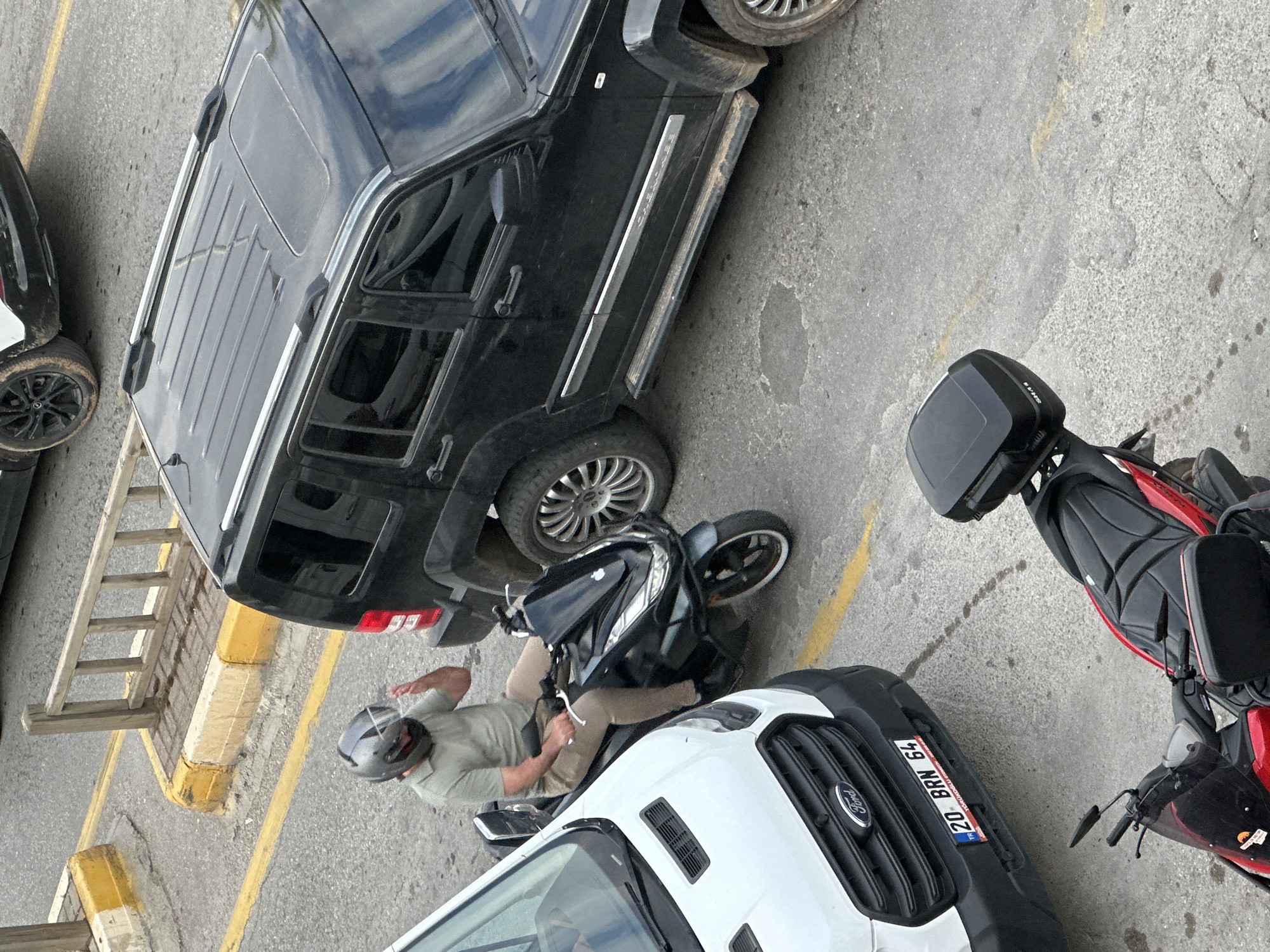 The scene depicts a street in Fethiye, Turkey. A black Jeep is parked adjacent to a white Ford Transit van. A person wearing a helmet and light green shirt is seated on a motorcycle between the two vehicles. Another red motorcycle is nearby. There is also a wooden ladder leaning against the curb. The van's license plate reads "20 BRN 64". The pavement is concrete.