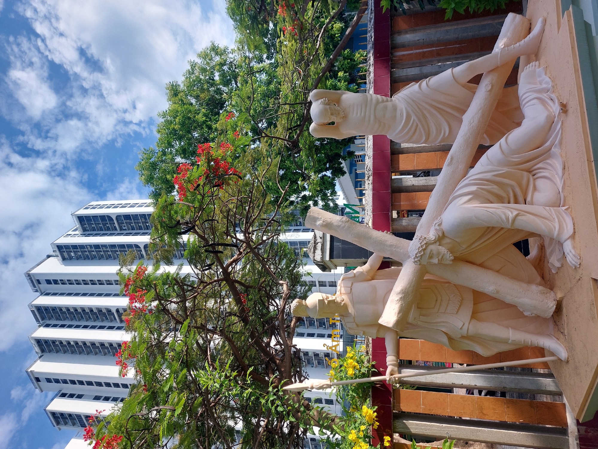 The image depicts a religious statue depicting Jesus carrying the cross, with a Roman soldier standing over him. The statue is placed outdoors in front of a row of modern high-rise buildings. Lush green trees with red flowers are visible in the foreground, framing the statue. The sky is blue with scattered white clouds. The overall scene suggests a public space within a city, possibly a park or a religious site. This scene is located at the Phat Quang Pagoda in Nha Trang, Vietnam.