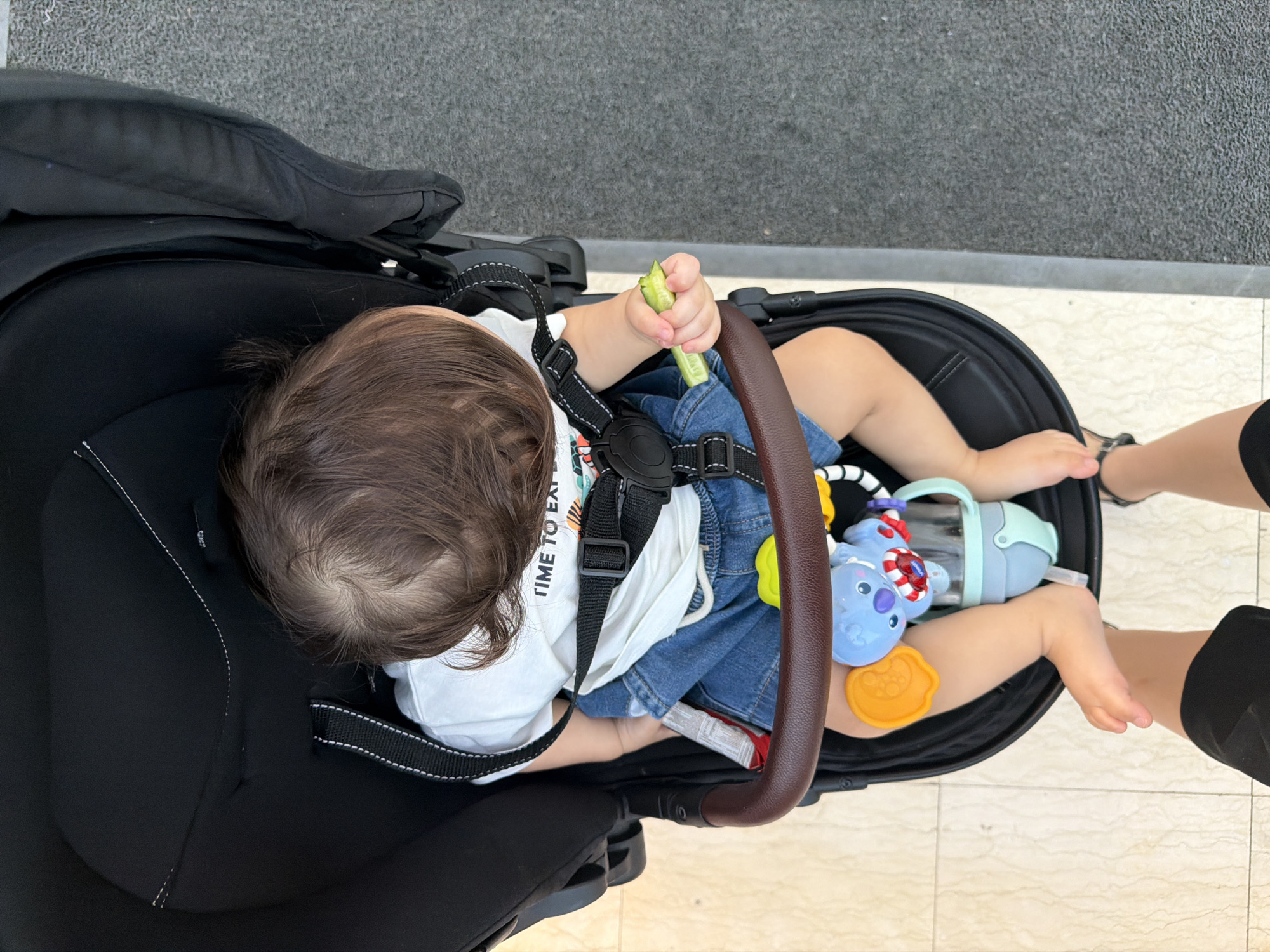 A high-angle, close-up shot shows a baby seated in a black stroller. The baby has brown hair and is wearing a white t-shirt with the text "TIME TO EXPLORE" and blue denim shorts. A black stroller harness is fastened around the baby. The baby's left hand holds a small green object, possibly a piece of food. A blue koala toy is attached to the stroller, along with a yellow ring toy and a sippy cup. The stroller is positioned on a tiled surface next to a dark gray, textured surface, possibly asphalt. The baby's feet are visible at the bottom of the frame, with the legs of an adult standing behind the stroller. The overall scene suggests a casual outing in an urban environment. It is not possible to determine the specific location within Sharm El Sheikh, Egypt, from the visual information provided.