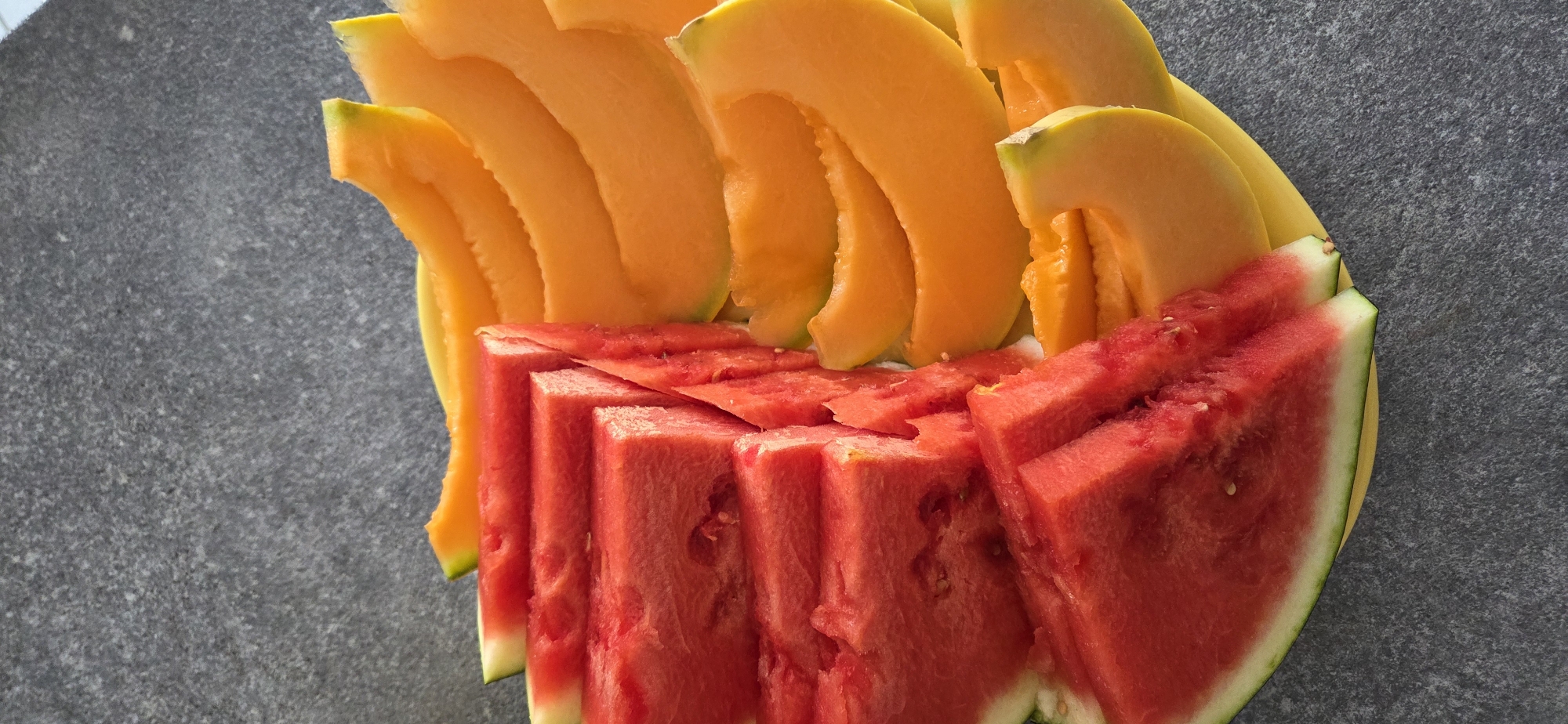 A close-up, vertical, eye-level shot shows sliced watermelon and cantaloupe arranged on a yellow plate. The watermelon slices, a vibrant red with white rinds, are stacked in a stepped pattern on the left side of the plate. The cantaloupe slices, a bright orange with a green-tinged rind, are fanned out on the right side. The plate rests on a dark gray, textured surface, possibly a countertop or table. The lighting suggests an indoor setting or an overcast outdoor environment, casting soft shadows. The image does not contain any specific clues to pinpoint its location within Lignano Sabbiadoro, Lignano, Italy.