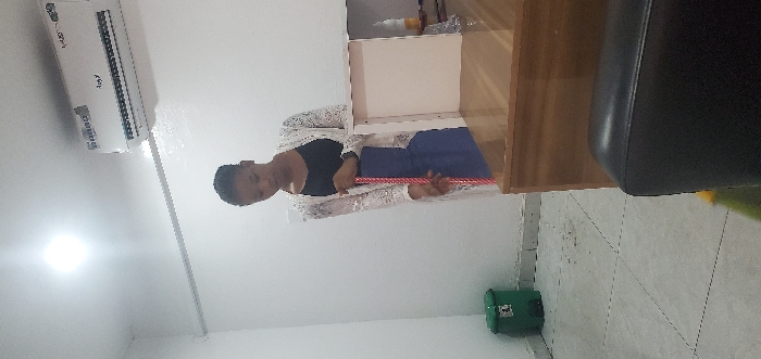 An indoor scene in an unidentified commercial or office space in Nigeria depicts a female individual. The specific street location is not discernible from the image.
A female with dark skin and short dark hair stands centrally, facing slightly to her left and looking downwards. She wears a black top, a blue skirt or dress, and a white, lace-patterned long cardigan. She holds a red and white striped pole, likely a broom or mop handle, vertically with both hands.
The room has white walls and a light-colored tiled floor with visible grout lines and some dark particulate matter near the left wall. A bright square ceiling light fixture is present in the upper left. A white, wall-mounted "Royal" brand air conditioning unit is fixed to the upper right wall. A white electrical conduit runs horizontally along the upper left wall.
To the right, a brown wooden desk or counter holds an open white box with internal dividers. Partially visible behind this box is a bottle, appearing to contain a yellow liquid. In the lower left, a green plastic waste bin with a black lid and a white label rests against the wall. A section of a black upholstered chair is visible in the bottom right foreground.