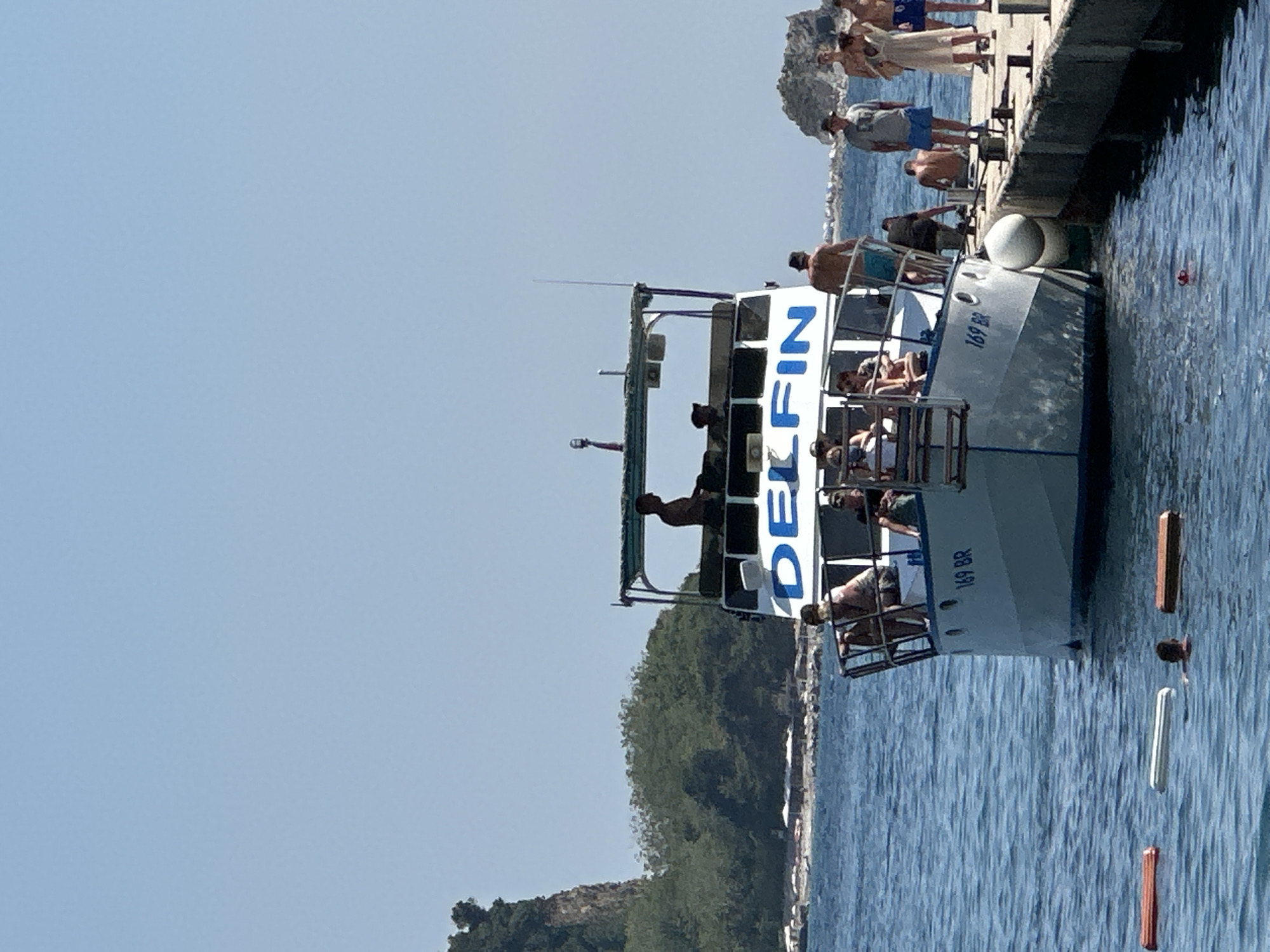 In a coastal scene off the coast of Budva, Montenegro, a boat named "DELFIN" is moored next to a concrete pier. The boat is a passenger vessel, with several people visible on its decks and the pier. The water is a calm, deep blue, reflecting the clear sky.
On the pier, a group of people are gathered, some standing and observing the boat. They are dressed in casual summer clothing, suggesting a warm, sunny day, likely midday given the bright light. One person is seen wearing a light-colored towel.
The "DELFIN" boat has a white hull with prominent blue lettering that reads "DELFIN" along its side. The boat also has a metal railing structure on its upper deck where passengers are seated, enjoying the view. Some individuals are leaning on the railing, and others are sitting. The boat is numbered "169 BR."
In the water near the boat, several buoys are floating, some wooden and some lighter in color. The background shows a rocky, tree-covered coastline, characteristic of the Adriatic region. The overall atmosphere appears relaxed and leisurely, consistent with a tourist activity or a day at the seaside.
