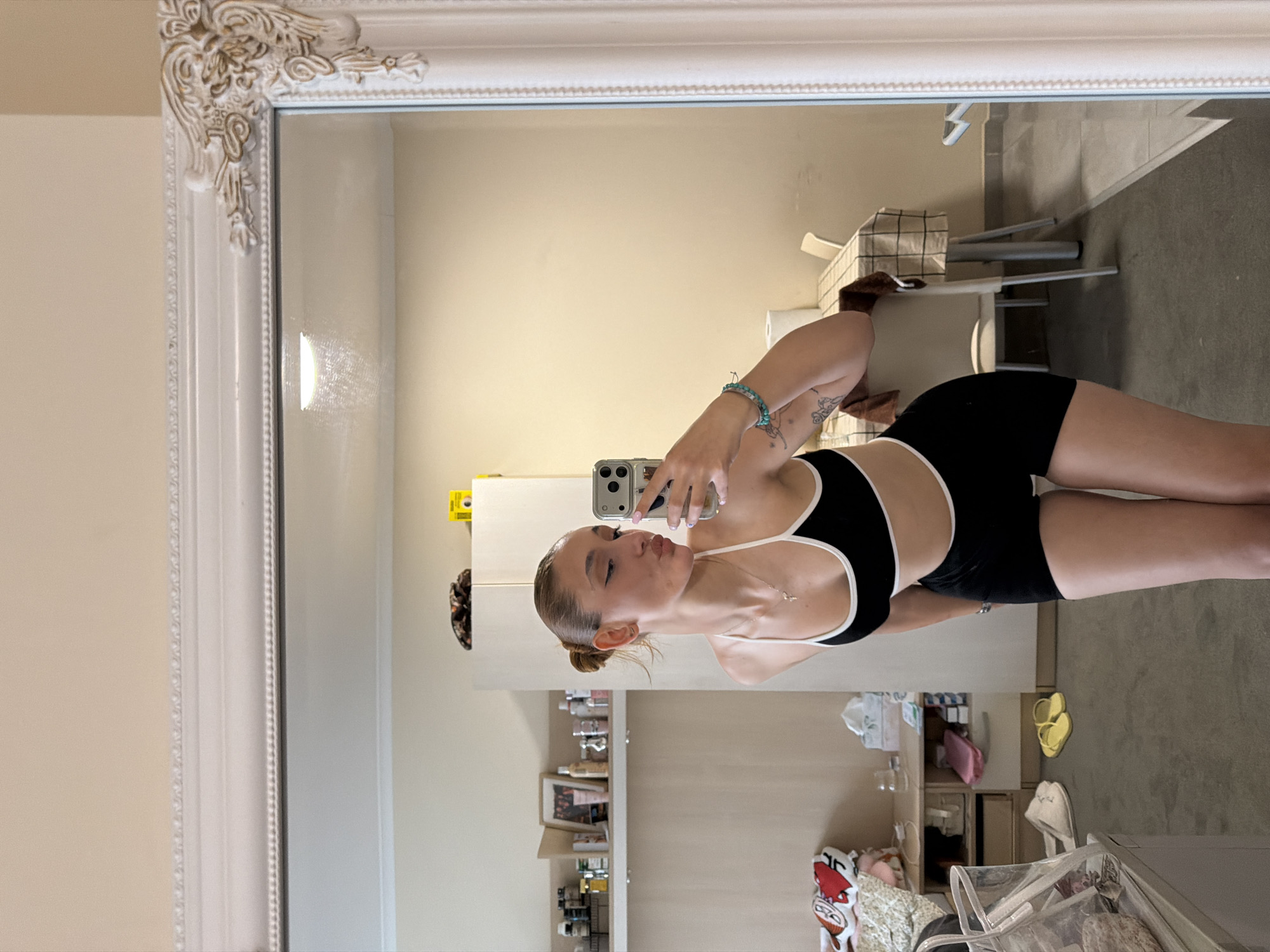 A young woman is taking a mirror selfie indoors. She wears a black and white sports bra and black shorts. She holds a silver phone with her left hand. The background shows a room with beige walls, a white shelving unit stocked with various items, and a glimpse of a bathroom with a tiled floor and a toilet. A white ornate mirror frame surrounds the image. The image's location is not identifiable as Sofia, Bulgaria, based on the available visual information.
