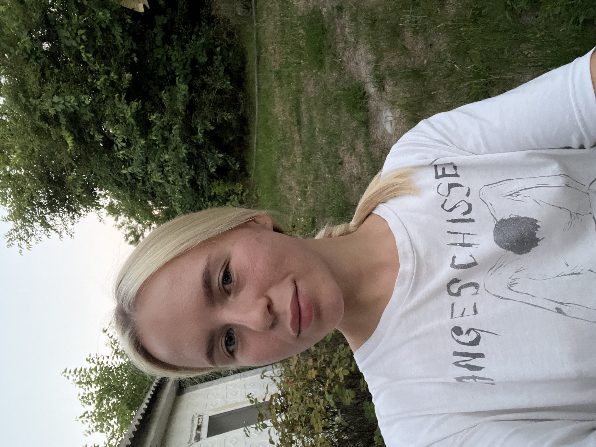 A young adult Caucasian female with blonde hair pulled into a ponytail, wearing a white t-shirt with a graphic print, is taking a selfie outdoors. She has blue eyes and fair skin. The background consists of a building with white siding and a grey roof on the left, green foliage, and a grassy area. The sky is overcast. The lighting suggests it is late afternoon or early evening. The image appears to have been taken in a rural or suburban setting.