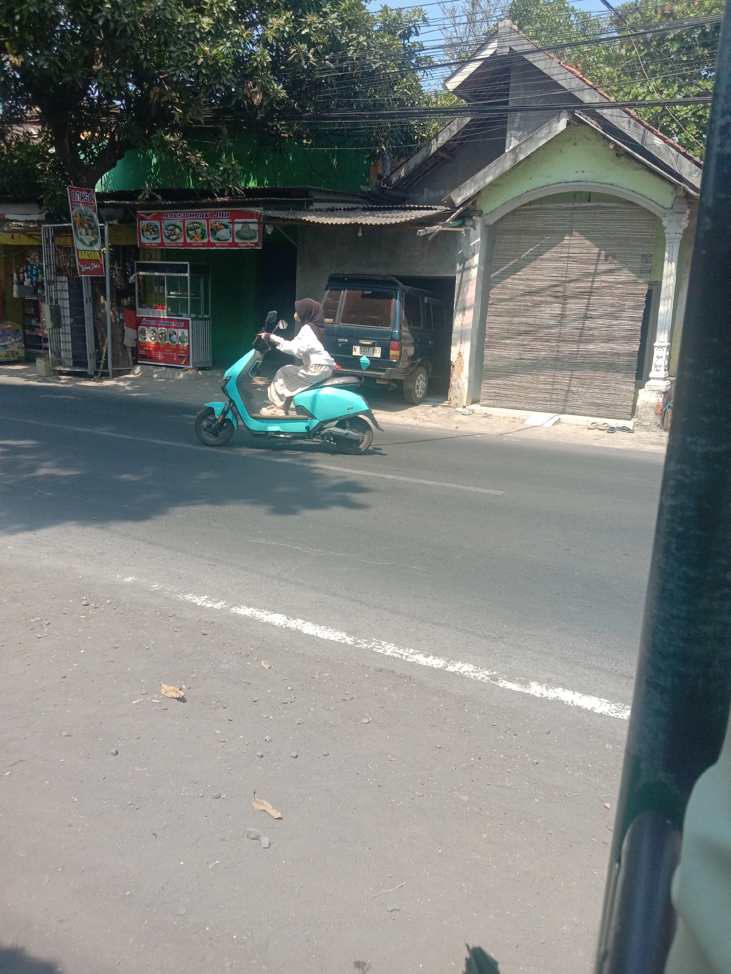A person wearing a white hijab and a long checkered skirt rides a turquoise electric scooter down a paved road in Karanganyar, Indonesia. The scooter is facing left. Behind the scooter, a dark green four-door SUV is parked. To the left of the scooter and SUV, a building with a green facade and a corrugated metal roof has signs advertising food, including "BAKSO" and "BUBUR AYAM." Further left, a tall, thin metal structure holds several items for sale. To the right of the SUV, a building features a light green arched entryway covered by a bamboo roller shutter. The road has a white dashed line painted down the center. Trees with dense green foliage overhang the scene. The ground on the side of the road is dusty and scattered with small stones and dry leaves. A shadow from the scooter falls on the road to its left. The scene is illuminated by bright sunlight, suggesting it is daytime.