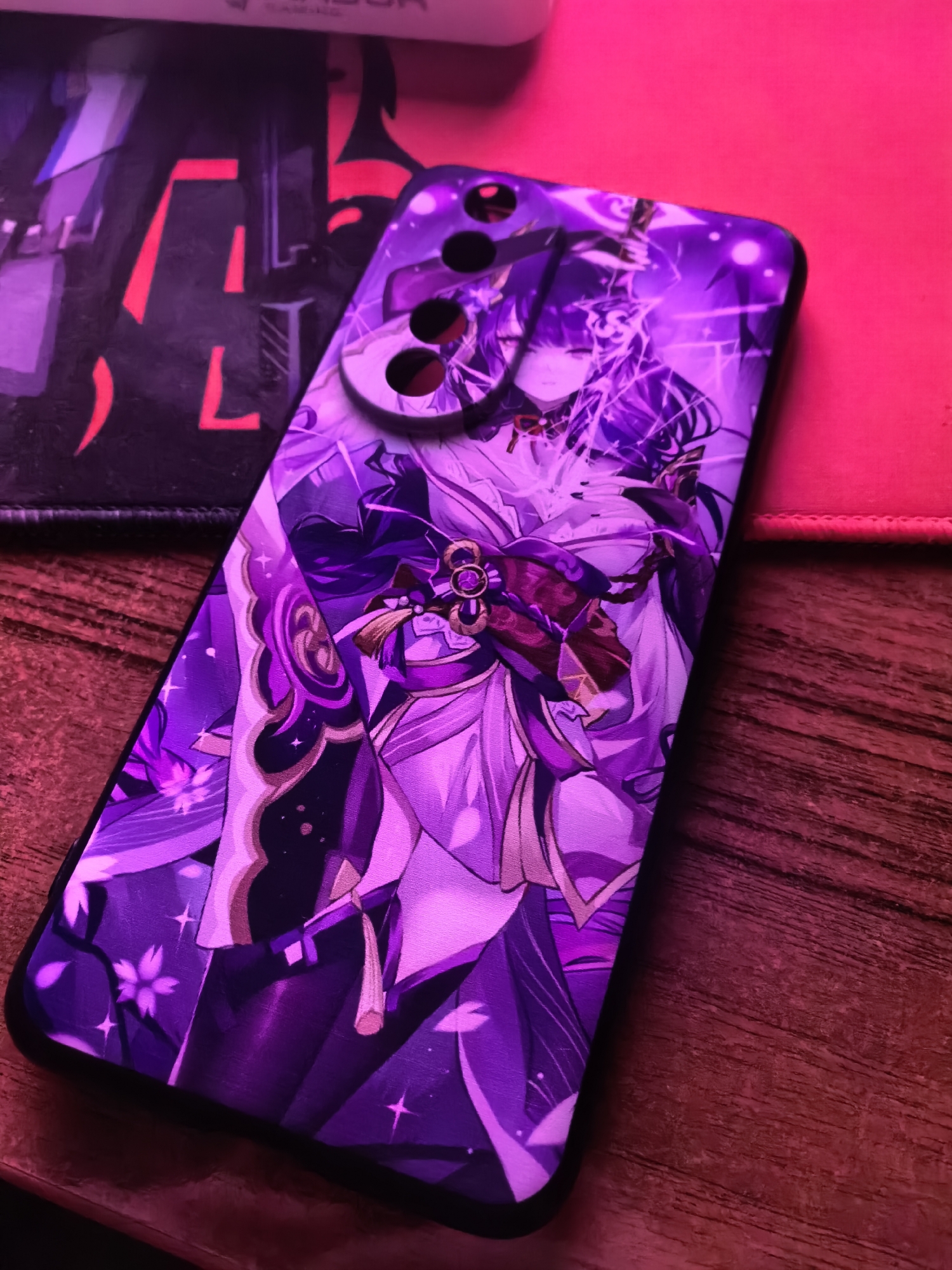 An anime-style image of a woman with purple hair and clothing is displayed on the back of a smartphone. The phone is placed on a wooden surface, with a black mousepad and a pink object in the background. The scene is illuminated by purple and pink lighting.